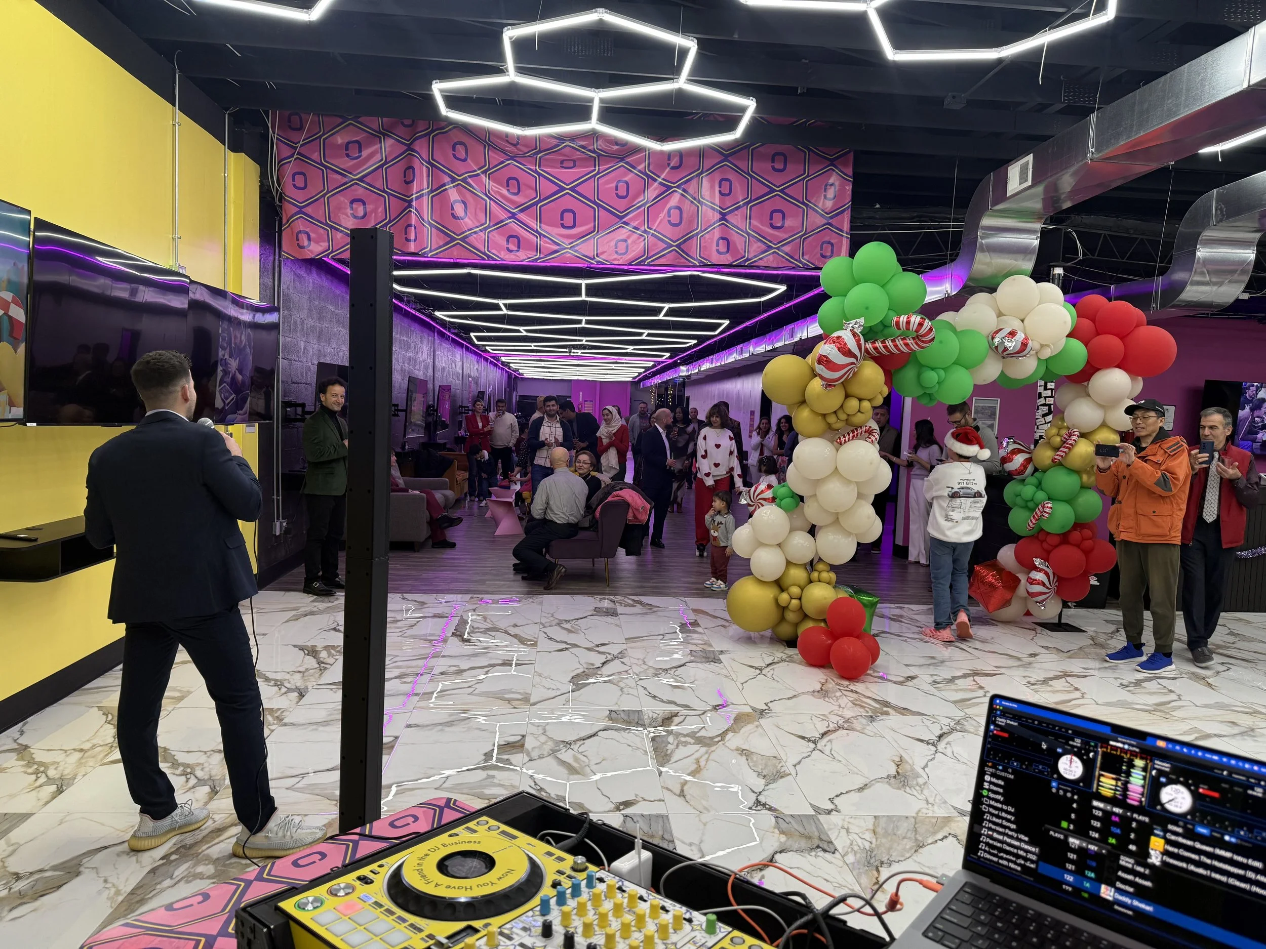 People gathering at a party or event with colorful balloon decorations, a DJ setup, and a dance floor in a modern indoor venue with vibrant lighting.