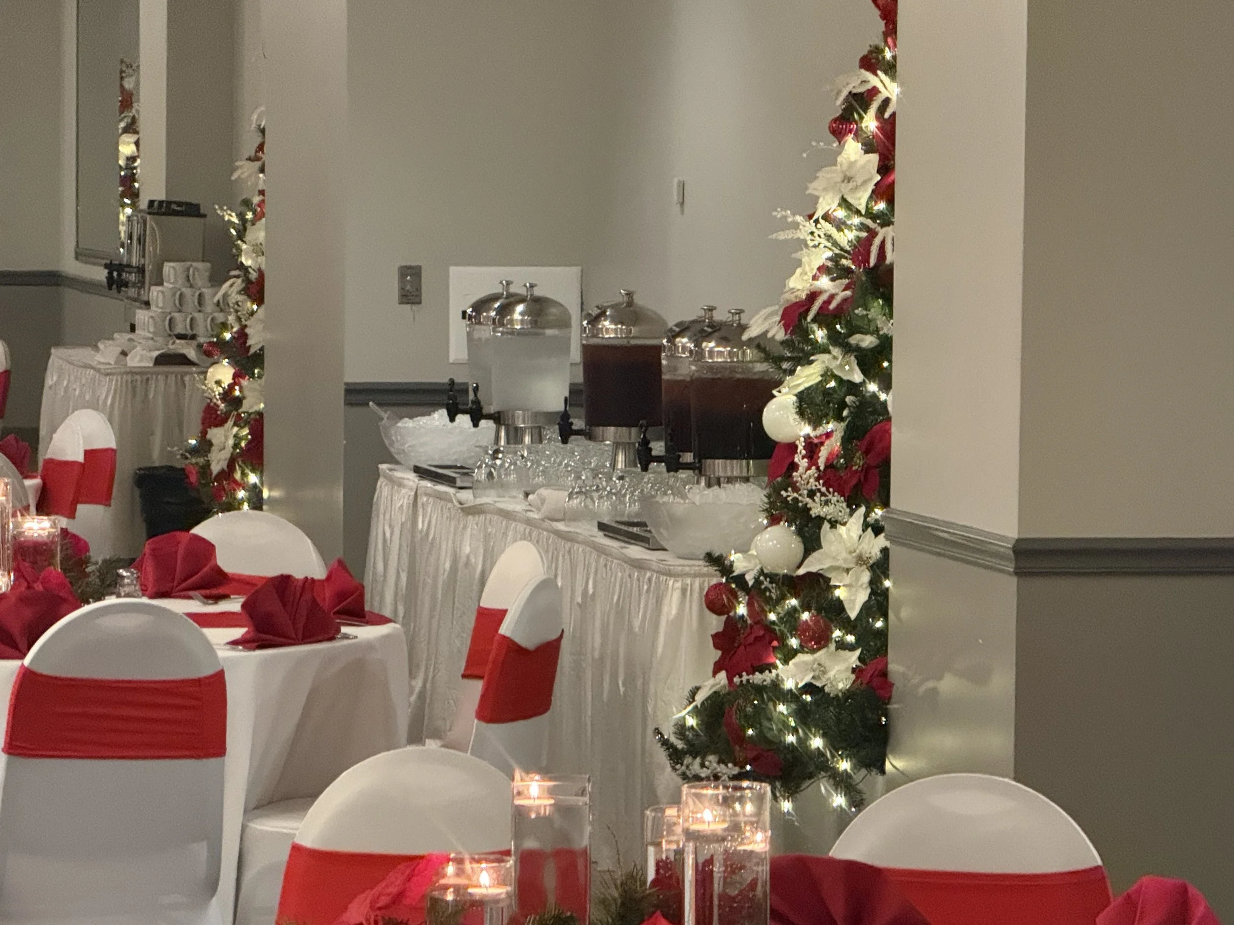 A festive banquet room decorated for Christmas with white chairs wrapped in red sashes, tables with red napkins, and a large floral garland with ornaments and lights. A table with beverage dispensers and glasses is visible in the background.