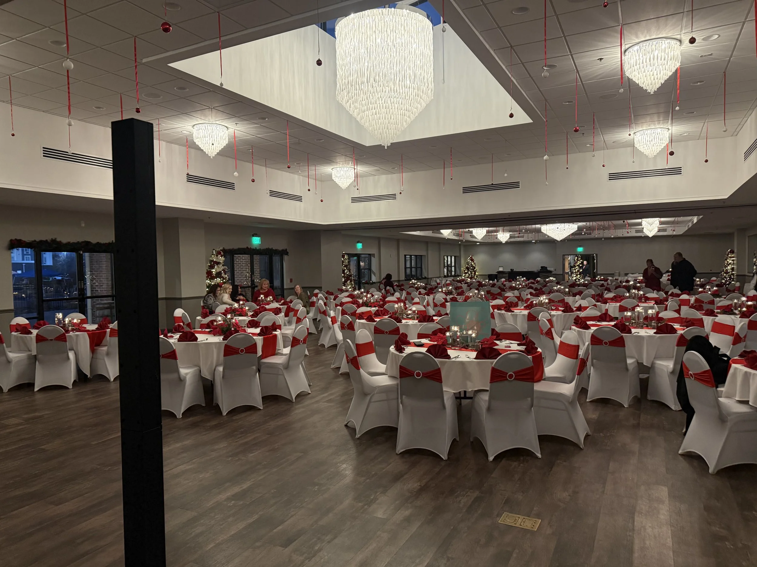 A banquet hall decorated for Christmas with round tables covered in white tablecloths, red napkins, and holiday centerpieces. The hall has a high ceiling with chandeliers, hanging ornaments, and windows along one side.