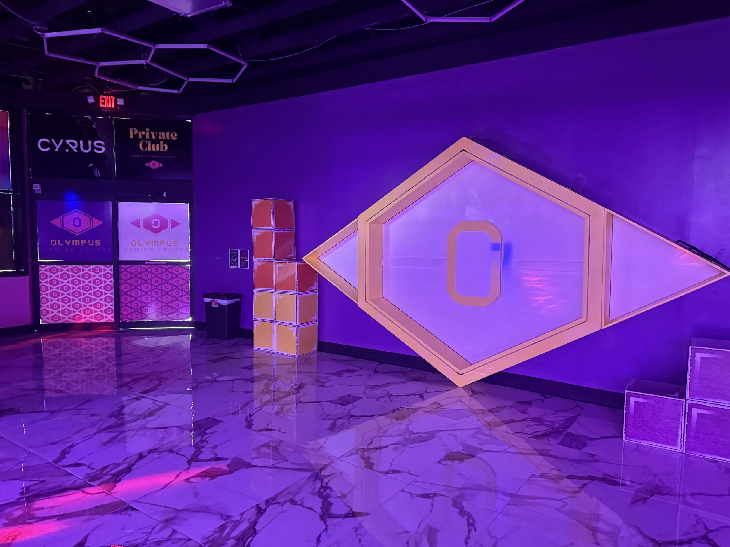 An interior of a gaming lounge with purple lighting, featuring a geometric wall decoration resembling a tilted diamond shape with a rectangular cutout, a stack of large yellow blocks, and illuminated signs that say 'CYRUS' and 'Private Club'.