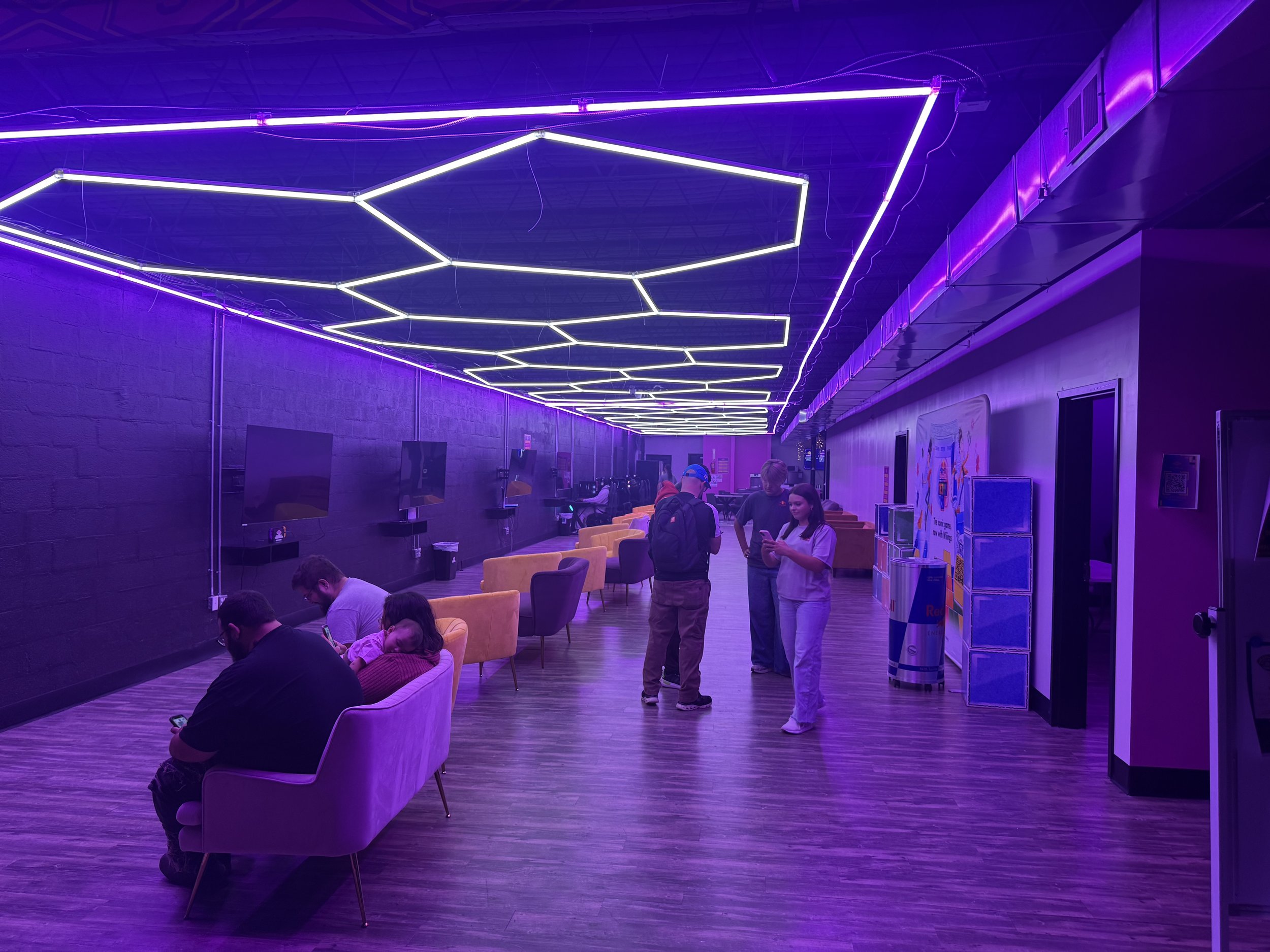 People sitting and standing in a room illuminated with purple and white neon lights, featuring chairs, televisions, and vending machines.