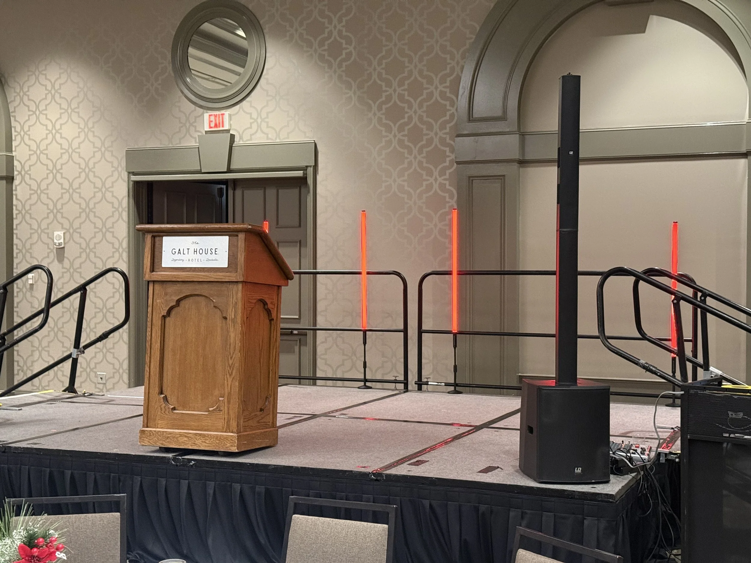 Empty stage with a wooden podium labeled 'Galt House' and a large black speaker with a microphone stand behind it. Red vertical lights are on the stage. The backdrop features patterned wallpaper and an arched doorway.