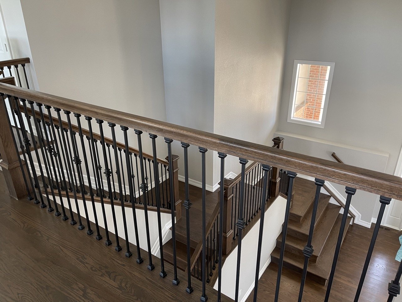 Interior view of a wooden staircase with black metal railings and a handrail, leading to the basement or lower level of a house. A small window with a view of a brick exterior wall.