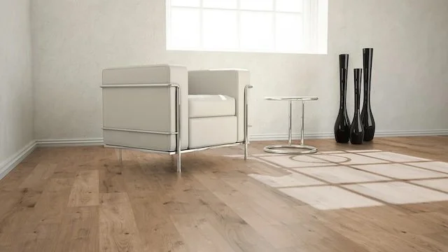 Minimalist living room corner with white armchair, small round side table, and three tall black vases on a wooden floor with sunlight casting shadows.
