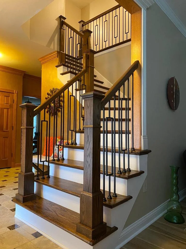 Interior view of a wooden staircase with black metal balusters in a home, featuring a landing and a top railing on the upper floor.
