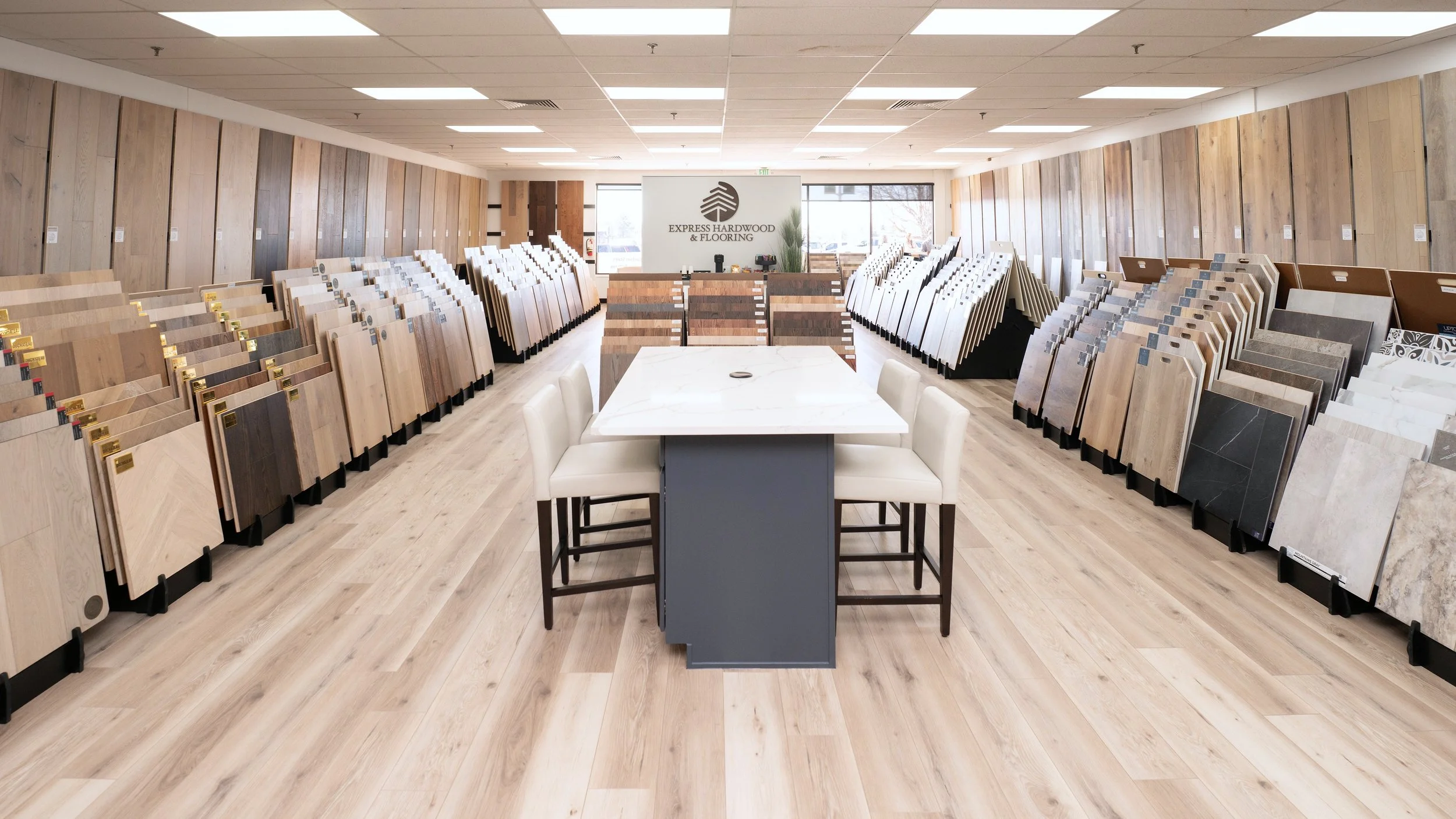 Interior of a flooring showroom with display samples of hardwood and tile flooring on the walls and racks, white table with chairs in the center, and a sign that reads 'Express Hardwood & Flooring' at the back.