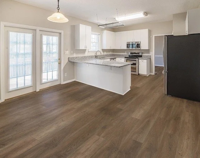 Empty kitchen with white cabinets, granite countertops, stainless steel appliances, a black refrigerator, and a wooden floor. Sliding glass doors lead outside.