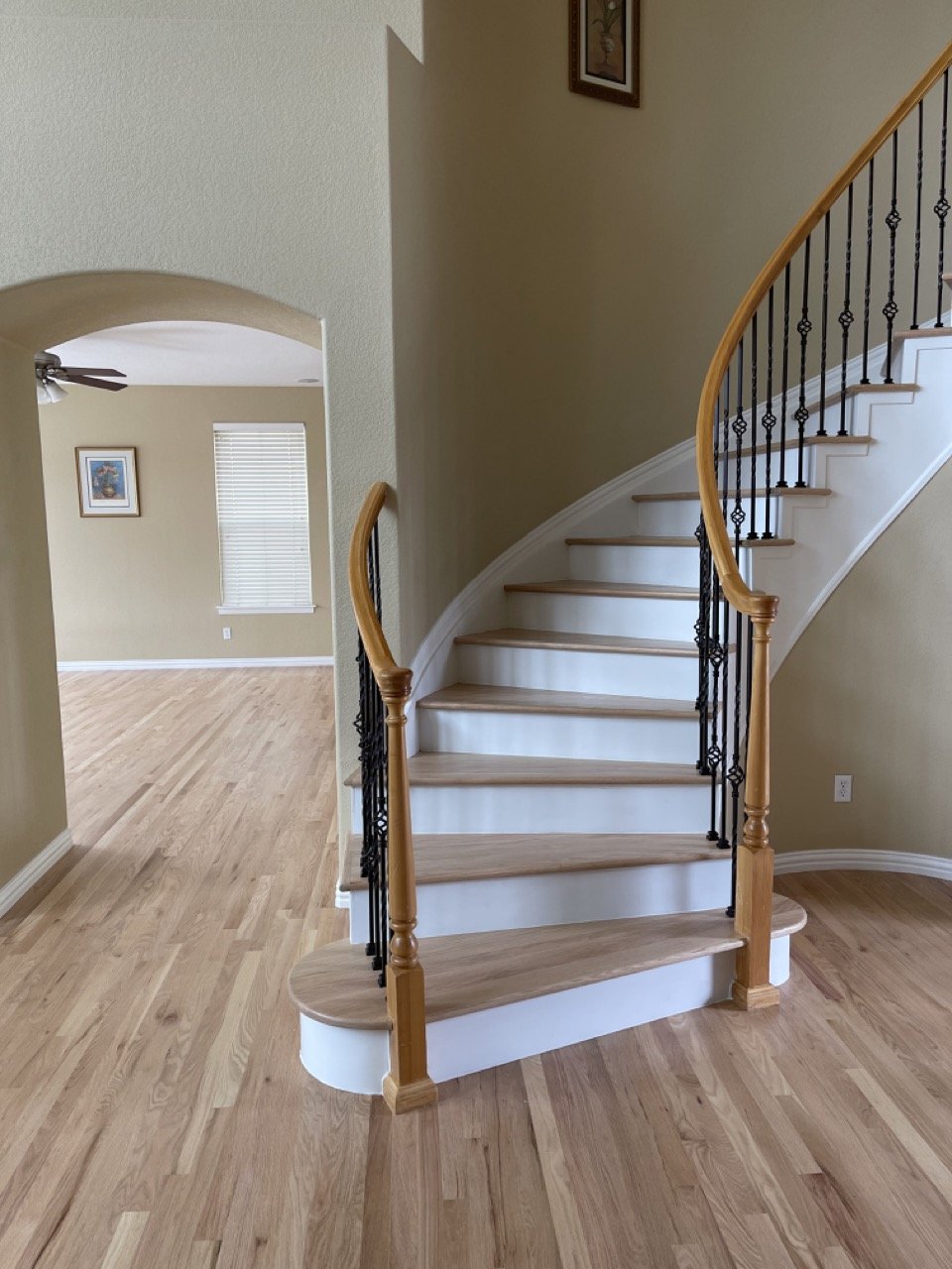 Interior of a home showing a curved staircase with wooden handrail and black metal balusters, wood flooring, and a nearby wall with a framed picture.