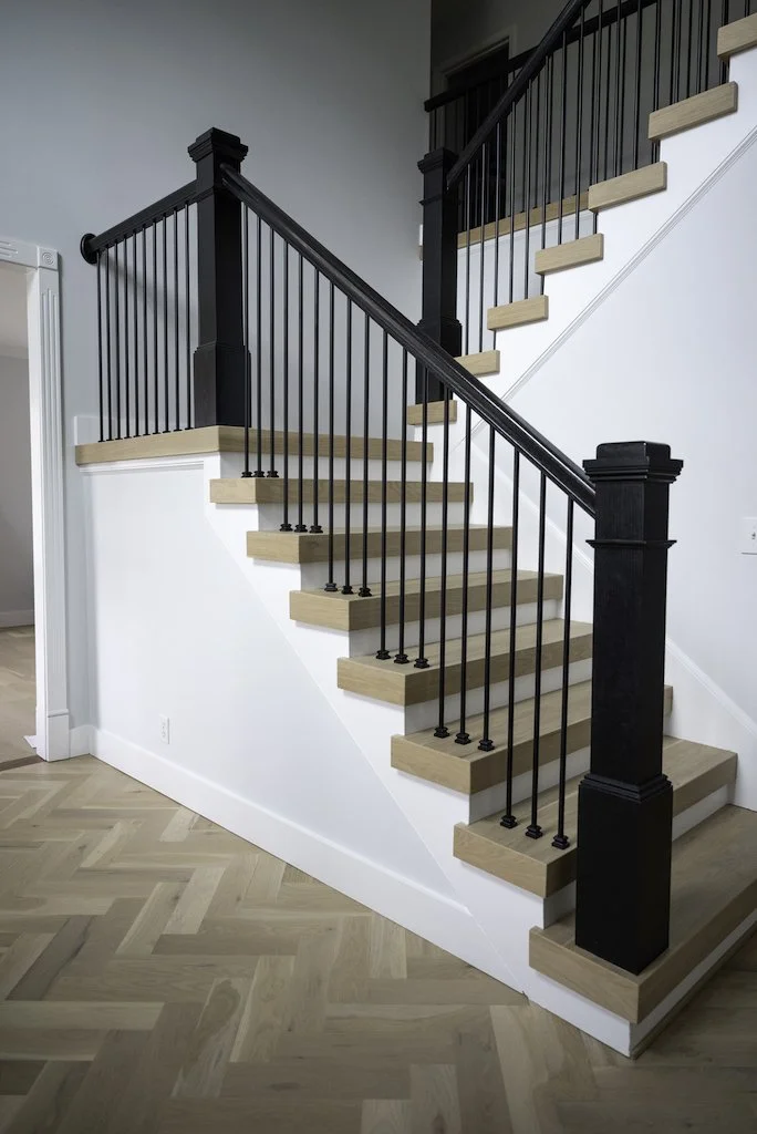Interior view of a staircase with light wood steps, black railing, and black newel posts, leading to an upper floor in a modern home.