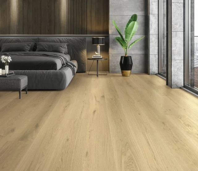 Modern bedroom with gray bed, black bedside lamp, small gray ottoman with white flowers, large green plant in black and gold vase, wood flooring, and glass wall with trees outside.