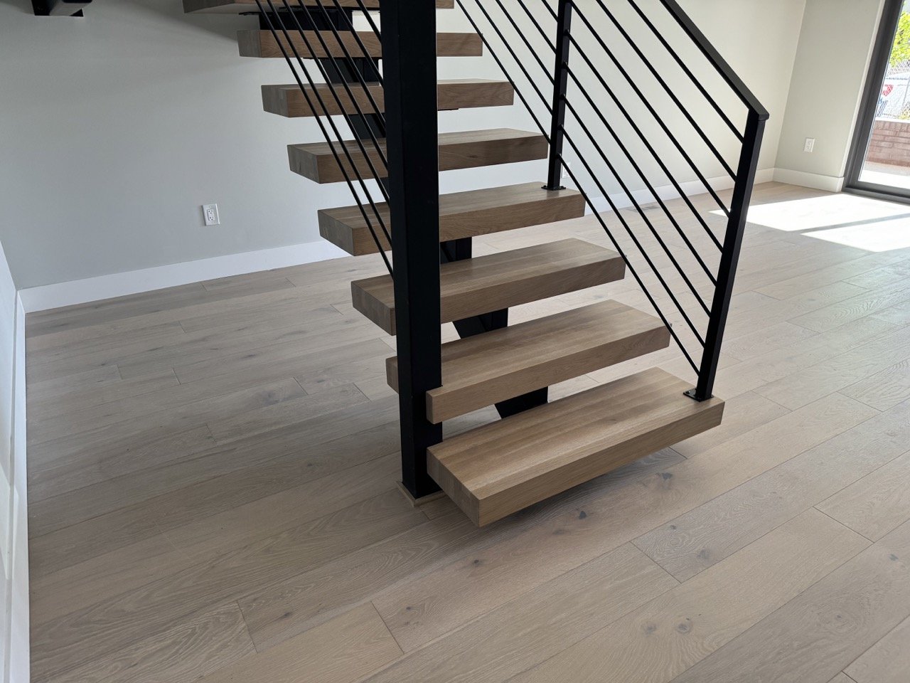 Interior view of a modern wooden staircase with black metal railings in a bright room with light-colored hardwood flooring and large windows.