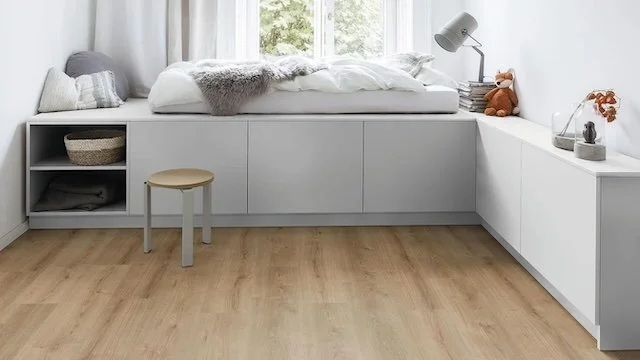 Minimalist bedroom with built-in white bed and storage, window with natural light, wooden floor, small stool, and decorative items.