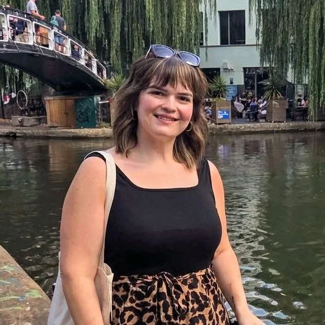A woman with shoulder-length brown hair and sunglasses on her head smiling at the camera near a river with a bridge and trees in the background.