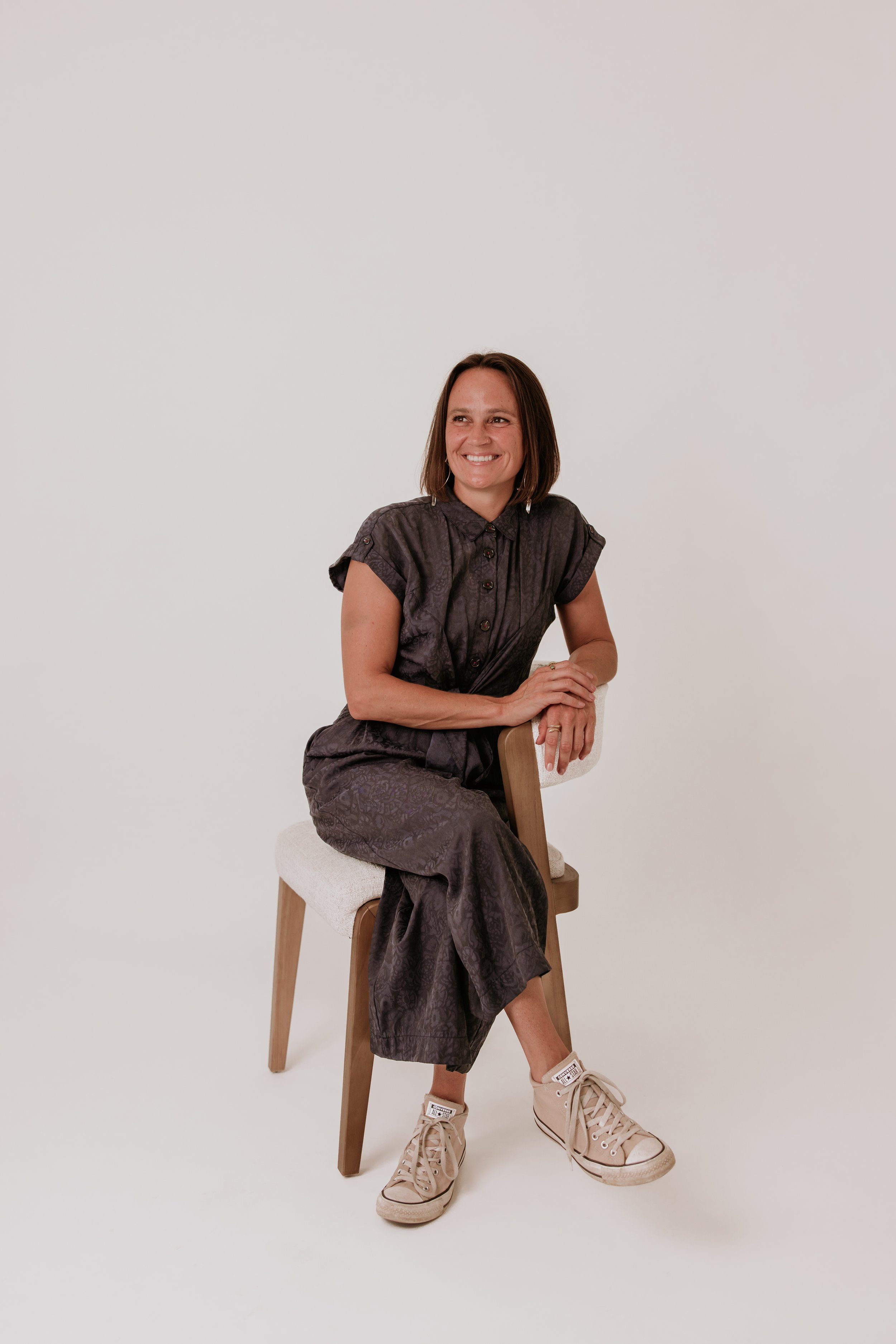 Woman sitting on a chair with a light-colored cushion, wearing a dark patterned outfit and beige high-top sneakers, smiling, with a plain white background.