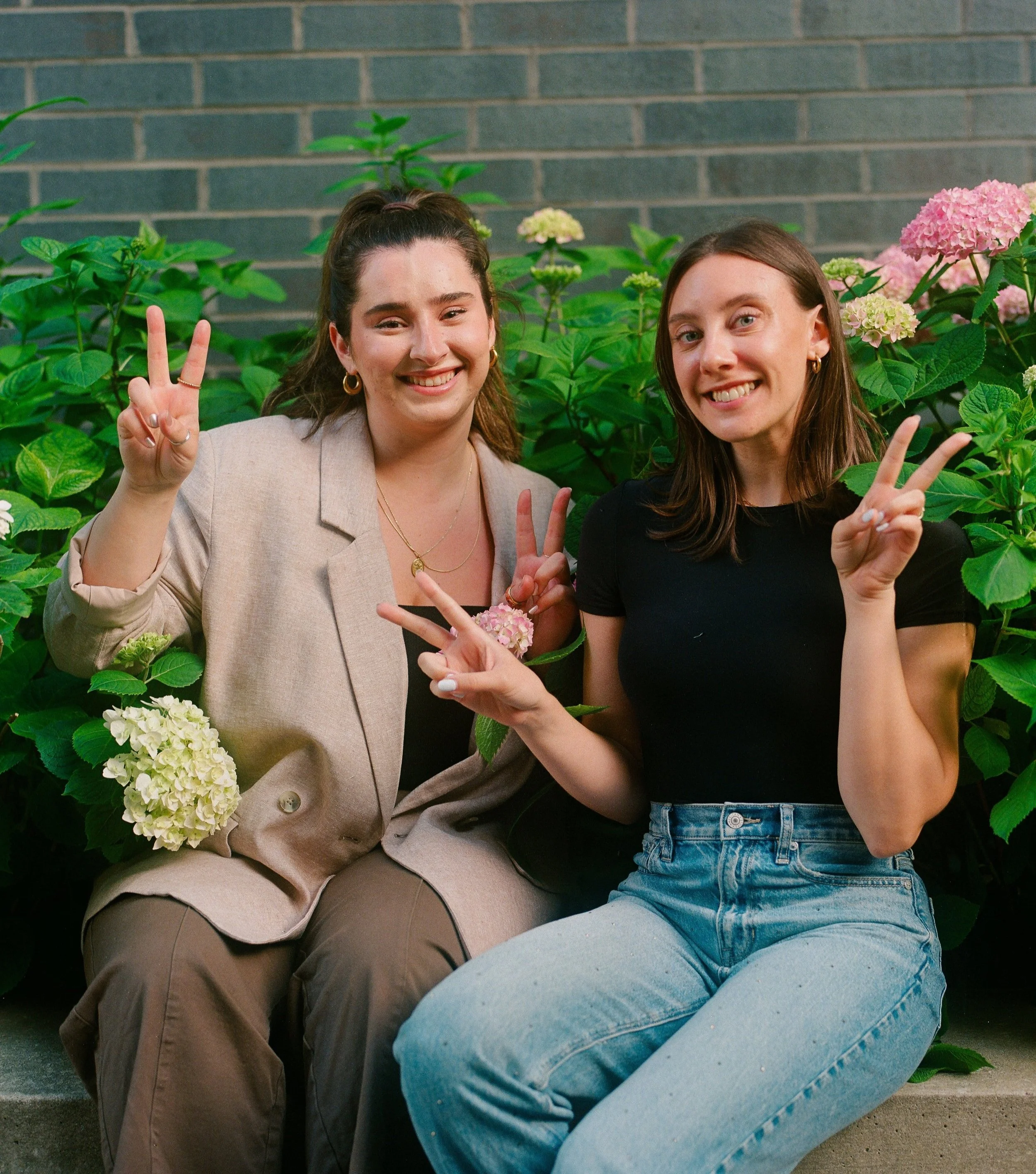 Two women sitting in front of blooming hydrangea plants, smiling, and making peace signs with their hands.