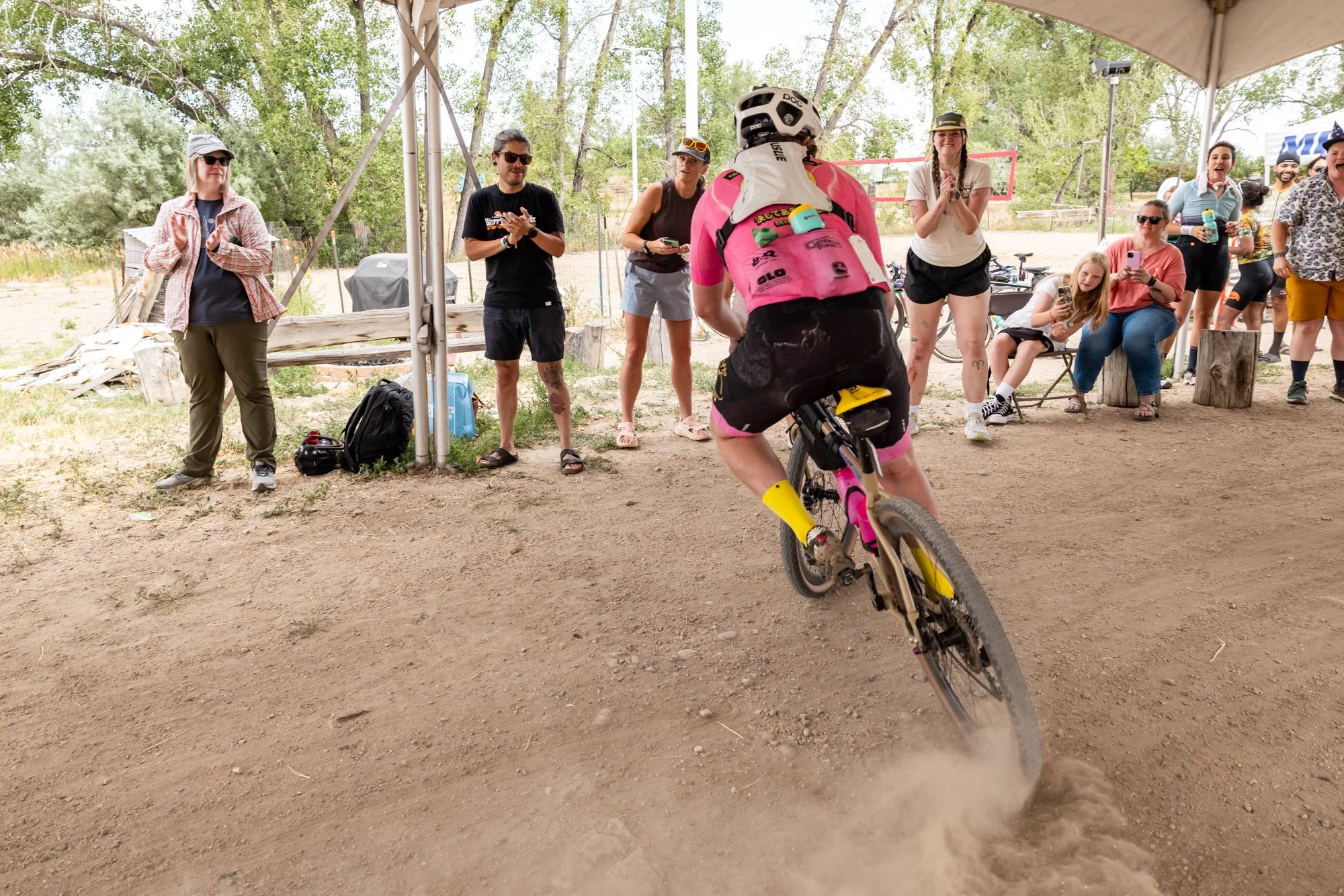 A group of people gathered outdoors under a canopy, watching a cyclist in pink gear riding on a dirt path. The audience is clapping and taking photos, with some seated on wooden blocks, and others standing, surrounded by trees.