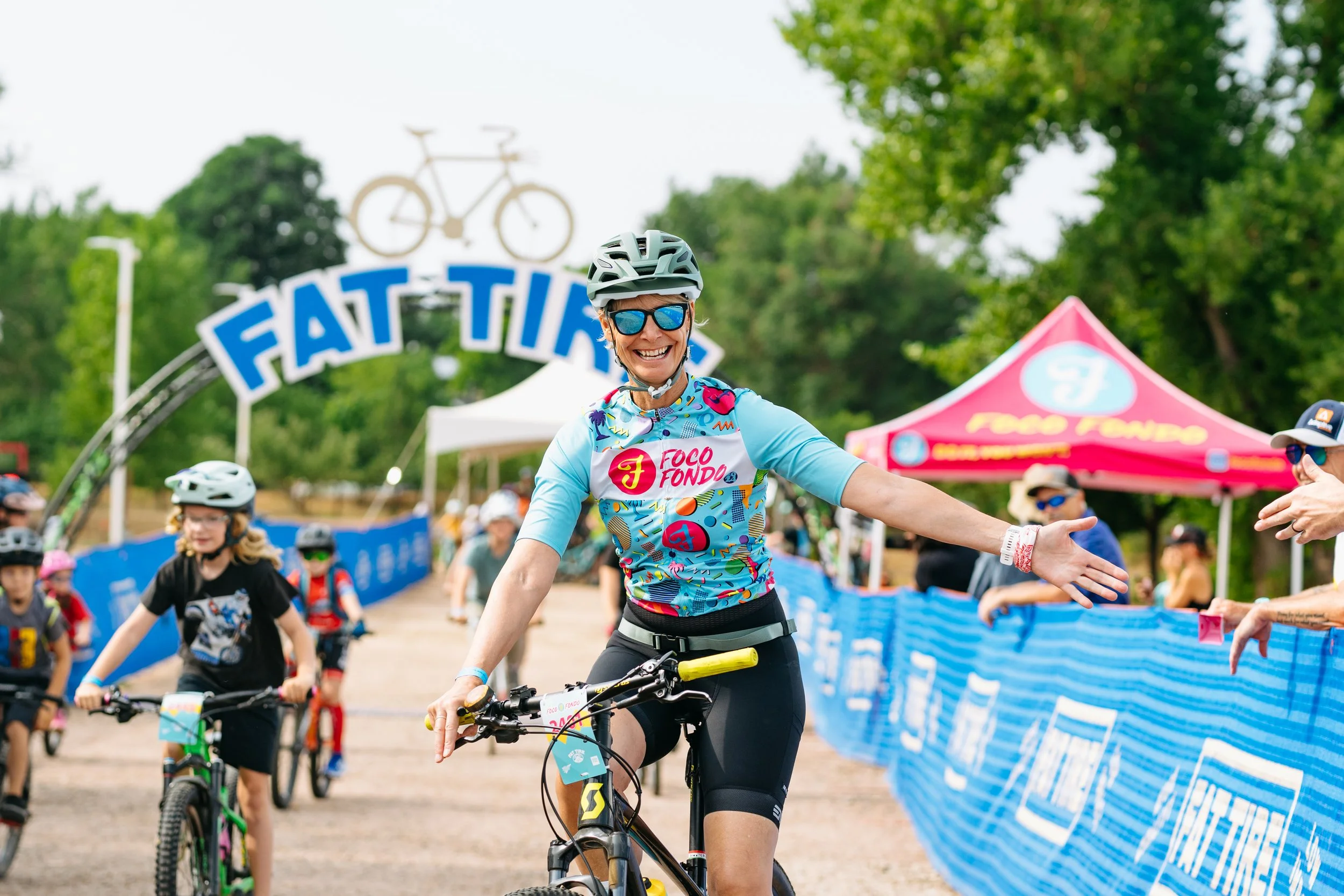 A man holding a woman in his arms in front of a backdrop with logos for Fat Tire and Foco Fondo, both in a cycling event.