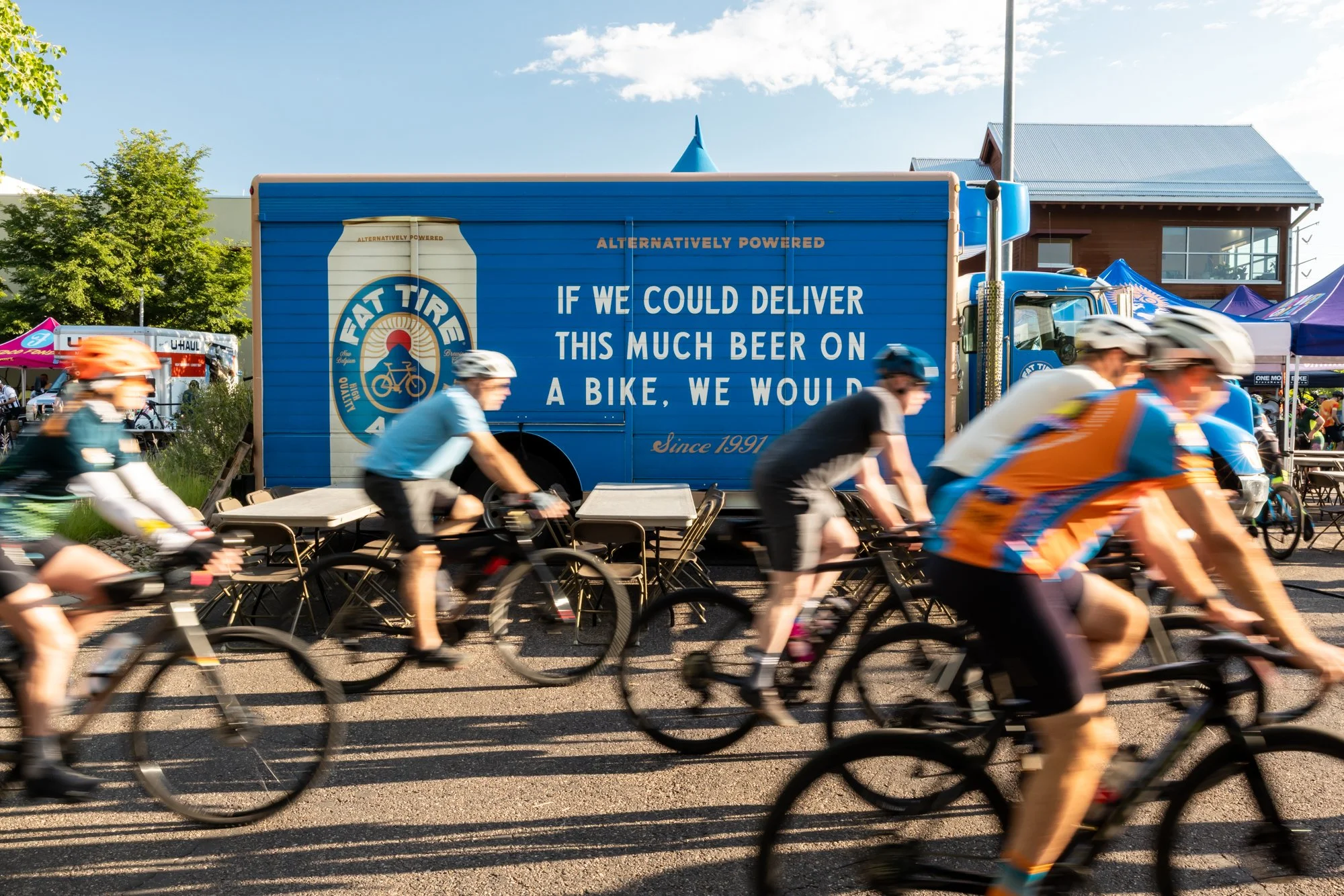 A group of cyclists riding past a Fat Tire truck at a festival. The truck displays an image of a beer can and a message about delivering beer on a bike.