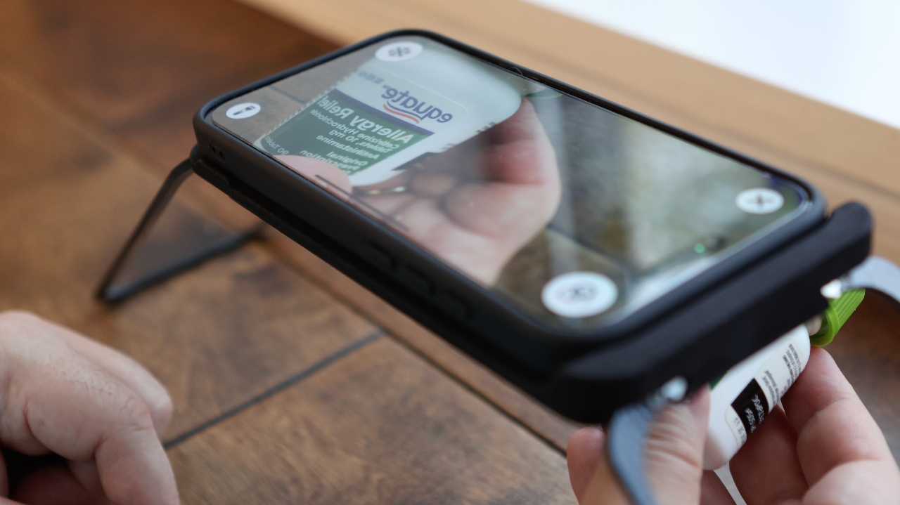 The Case for Vision app zooms in on the label of a medicine bottle held beneath an iPhone, making the small print readable on the screen.