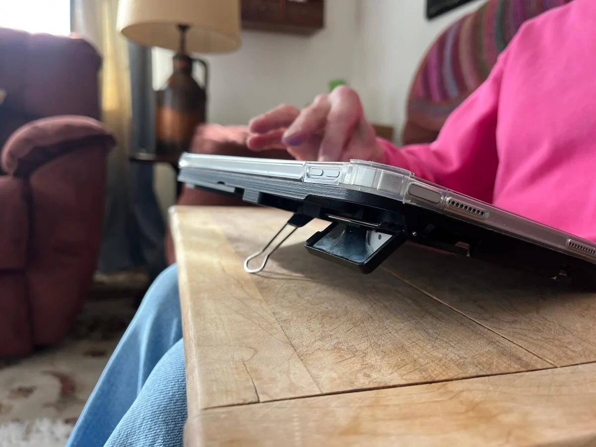 Side view of Case for Vision Solaris on a wooden table, showing the extended distance mirror and kickstand beneath the tablet while a woman in a pink sweatshirt uses the touchscreen.