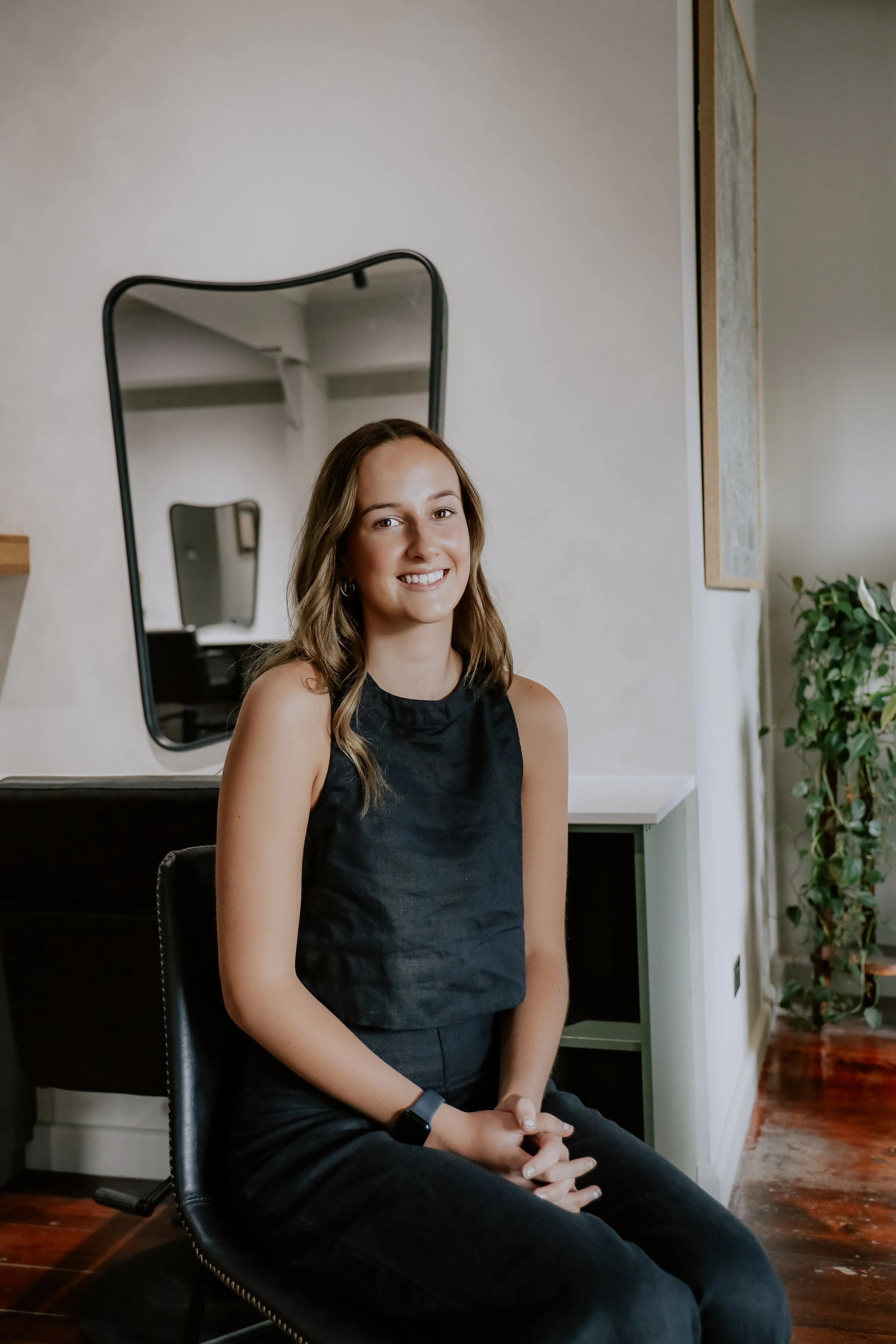 A young woman with long blonde hair smiling as she appears to be working or preparing something, wearing a black apron with white lettering, in an indoor setting.