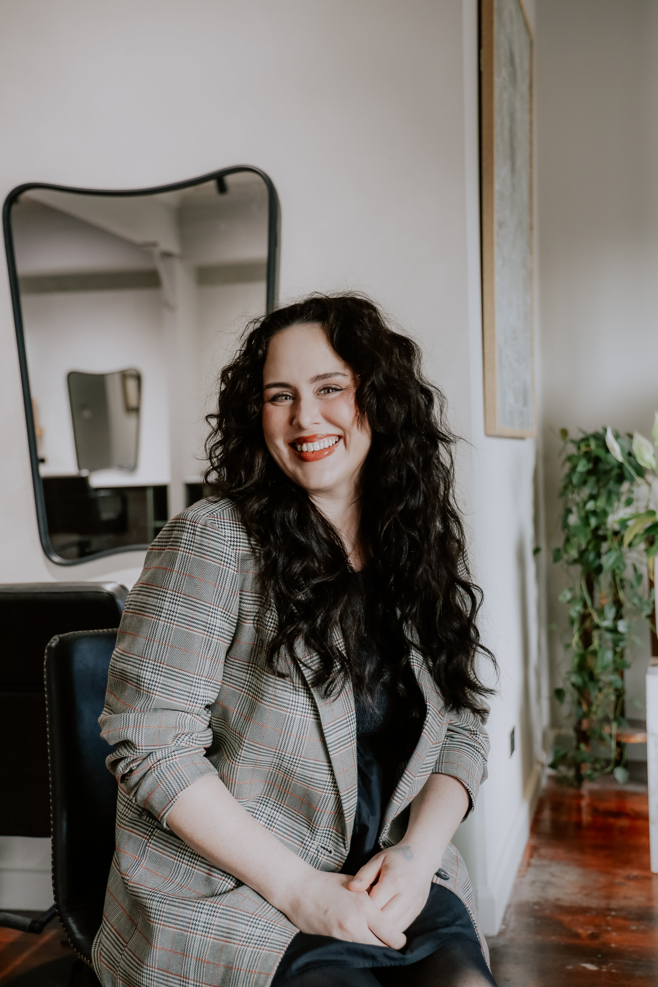 Smiling woman in checkered blazer in a store or salon setting, with shelves of products in the background.