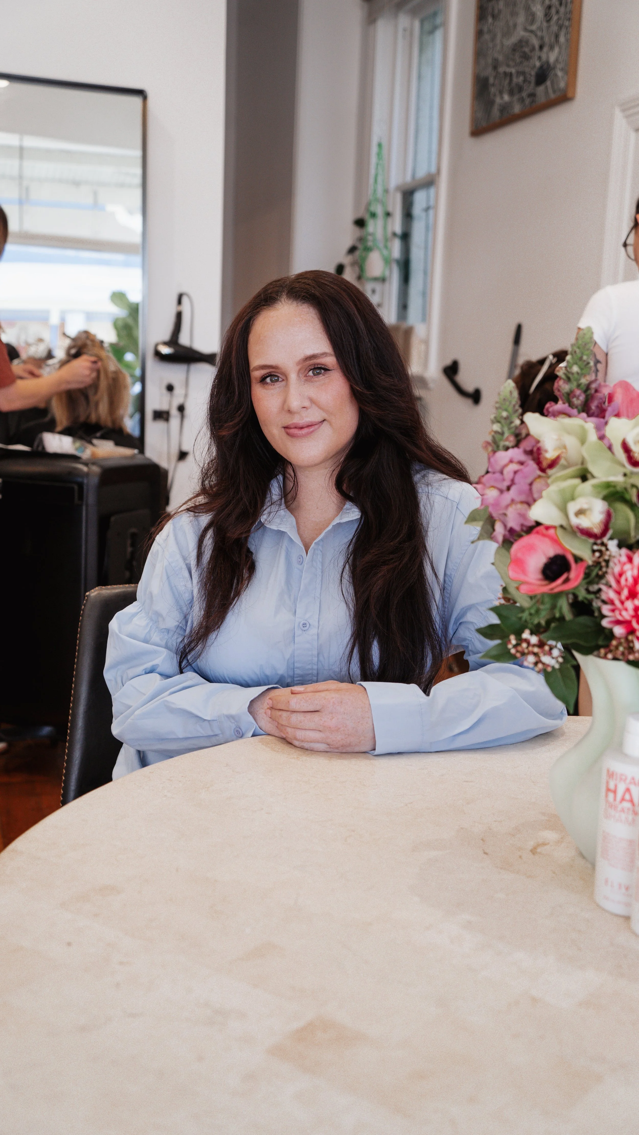 Smiling woman in checkered blazer in a store or salon setting, with shelves of products in the background.