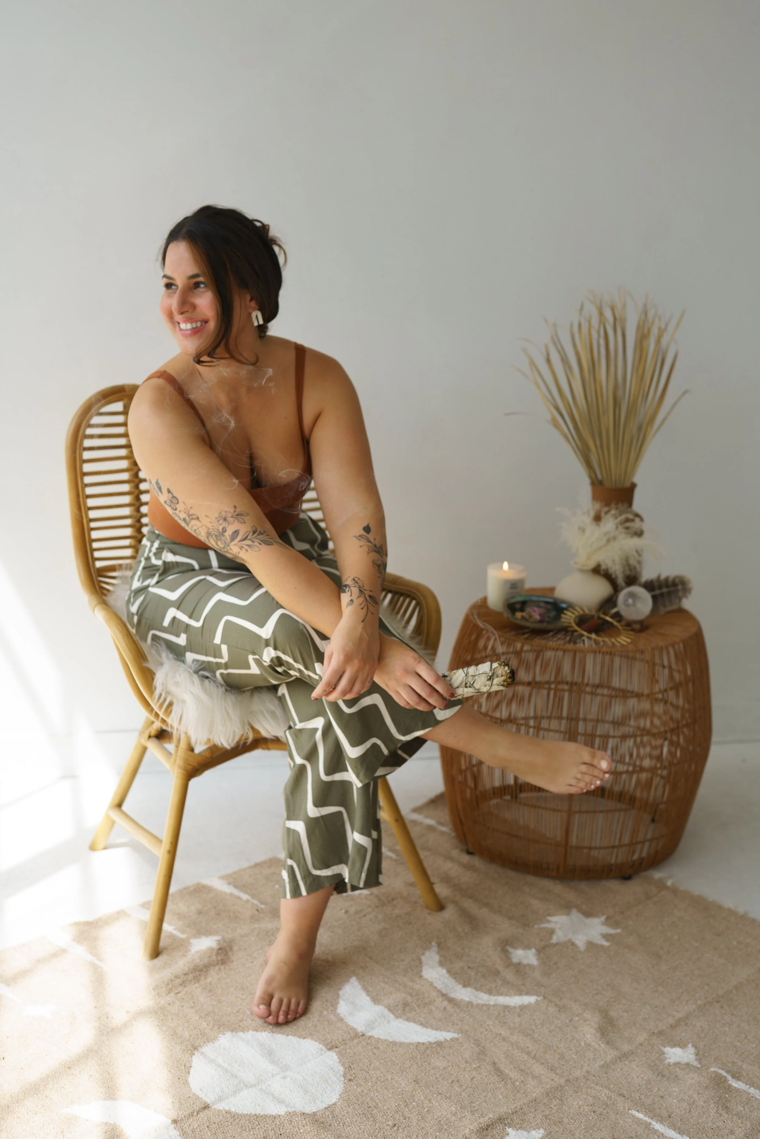 Stephanie Kamille sitting on a chair, holding sage, with a wicker table and a candle with spiritual items