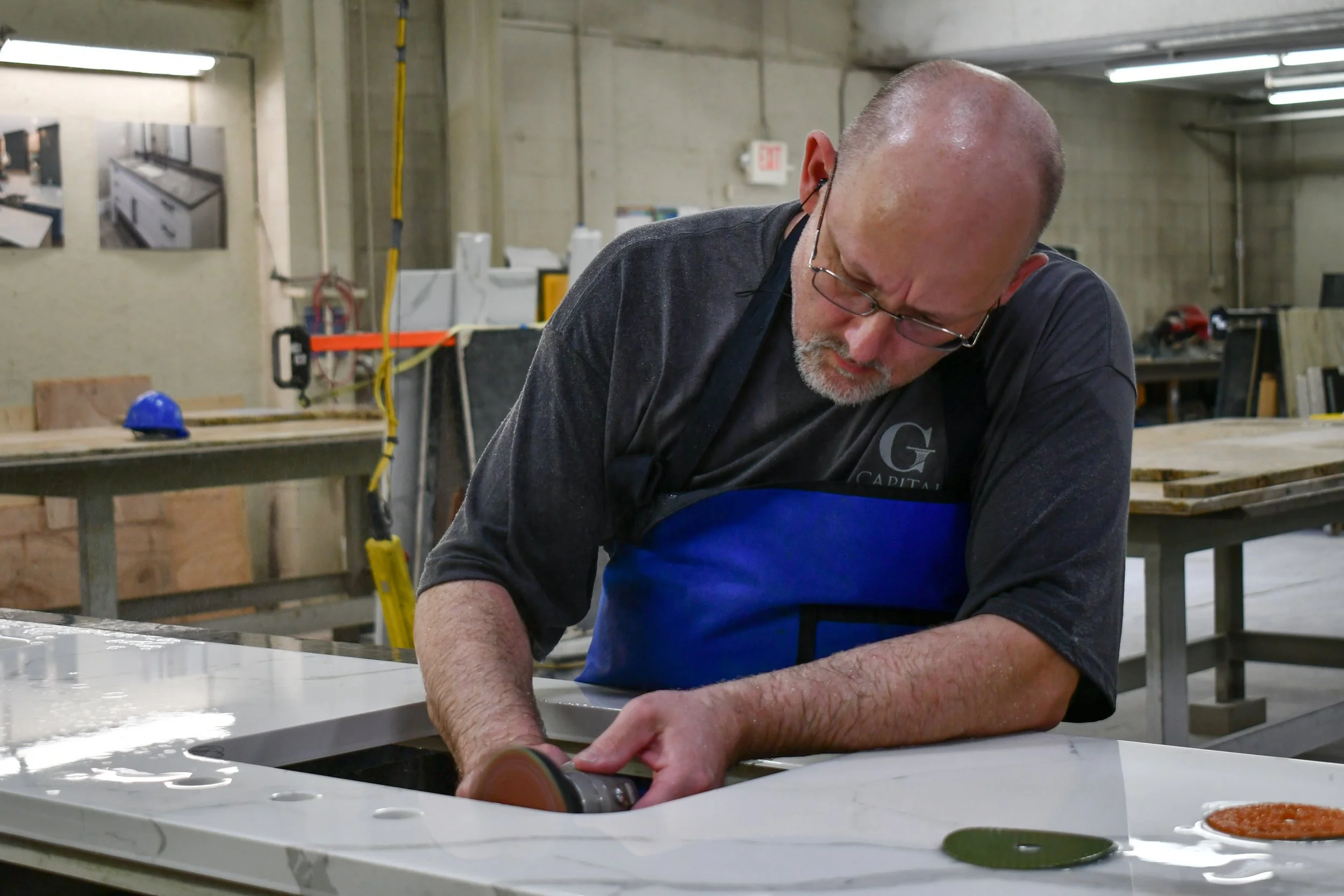A worker with headphones operating a robotic arm in a workshop setting. The workshop has tools on the wall and a granite slab on a workbench.