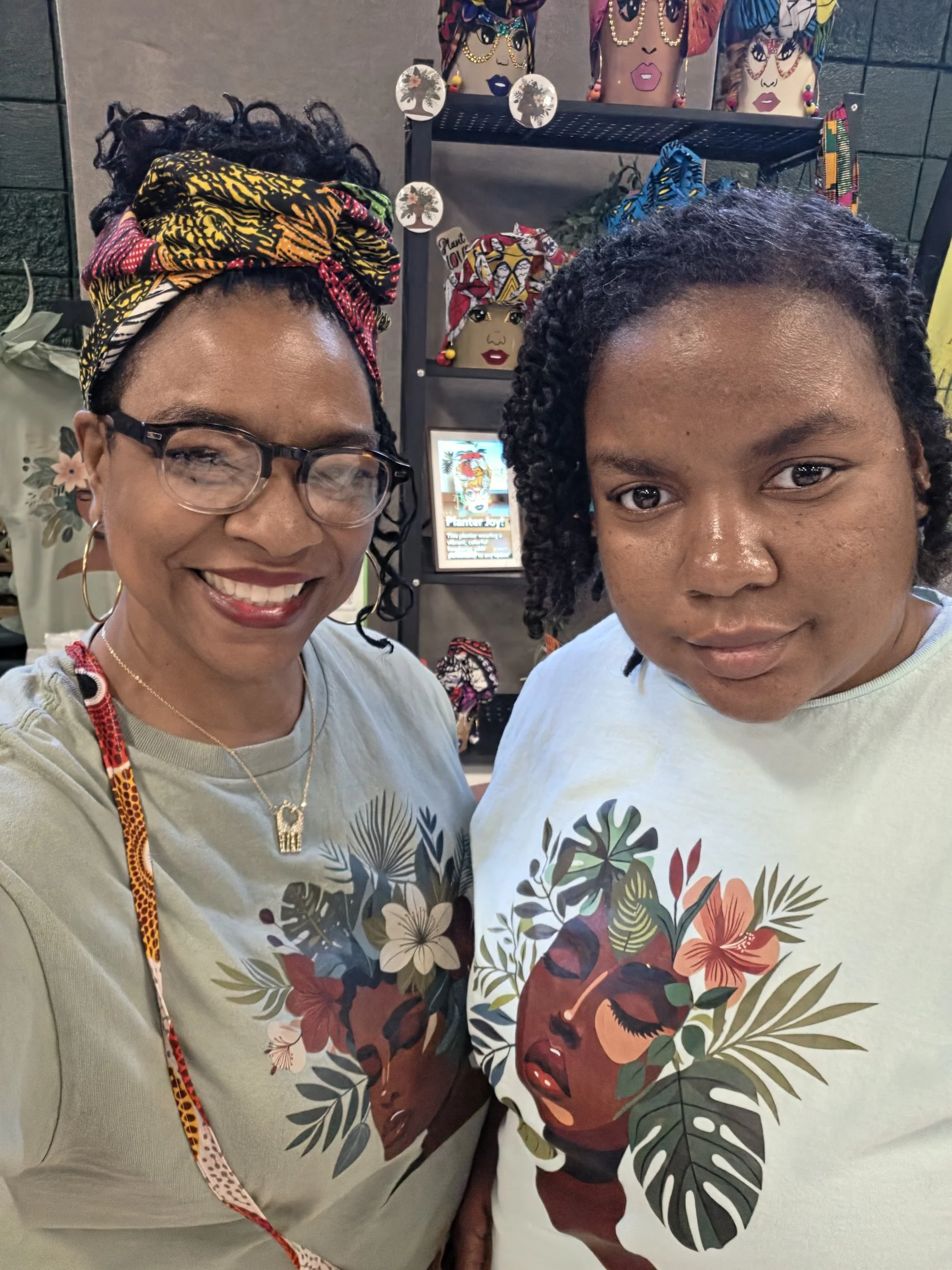 Two women taking a selfie in a store with colorful headscarves and handbags in the background. The woman on the left is wearing glasses, a headwrap, and smiling. The woman on the right has curly hair and is not smiling.