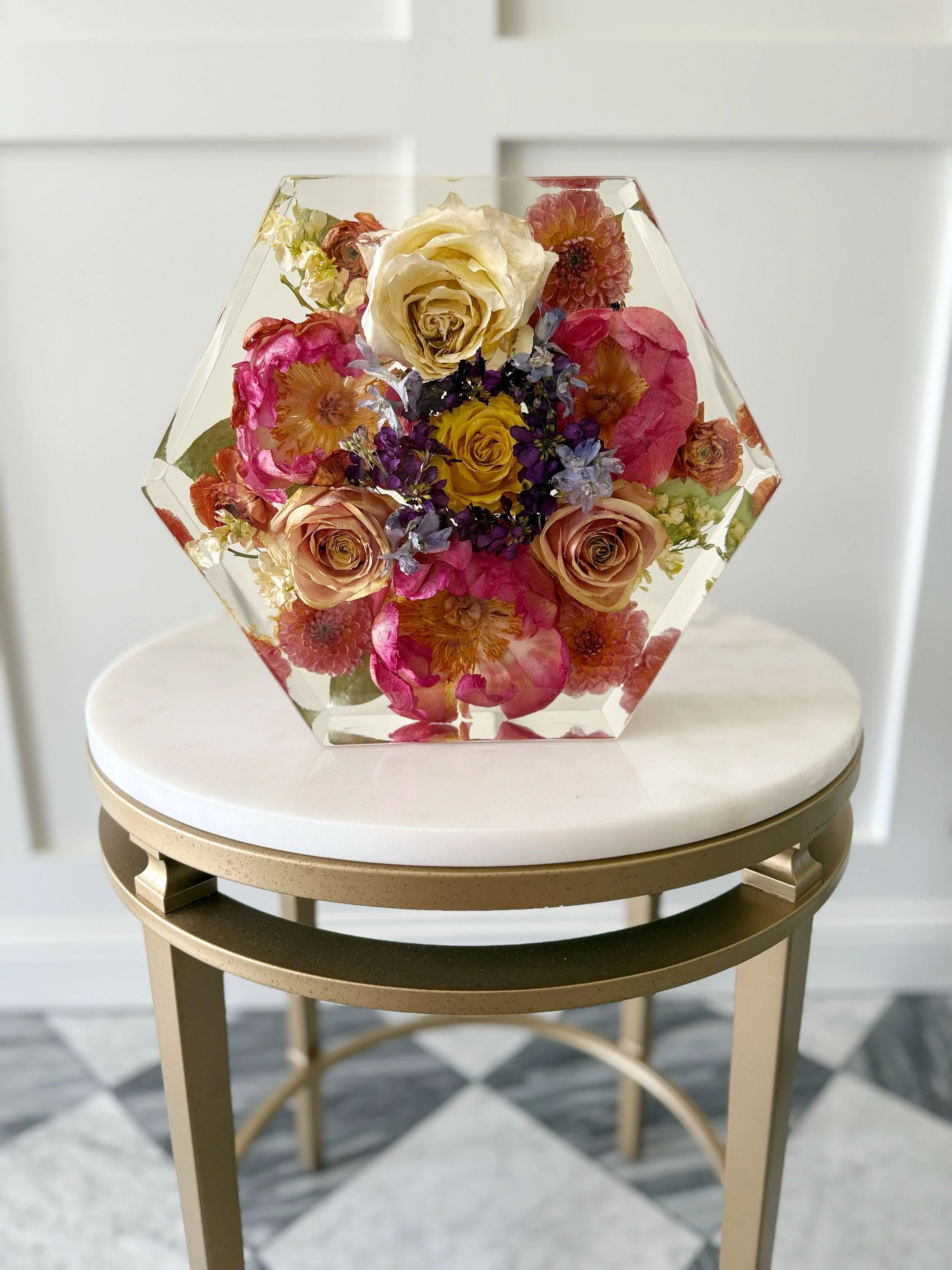 A glass hexagonal vase filled with various colorful flowers, including roses, dahlias, and other blossoms, placed on a small round table with a white marble top.