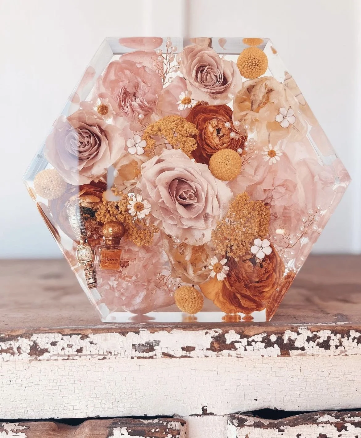 A hexagonal glass box filled with pink and orange roses, small white daisies, and yellow ball-shaped flowers, placed on a distressed white wooden surface.