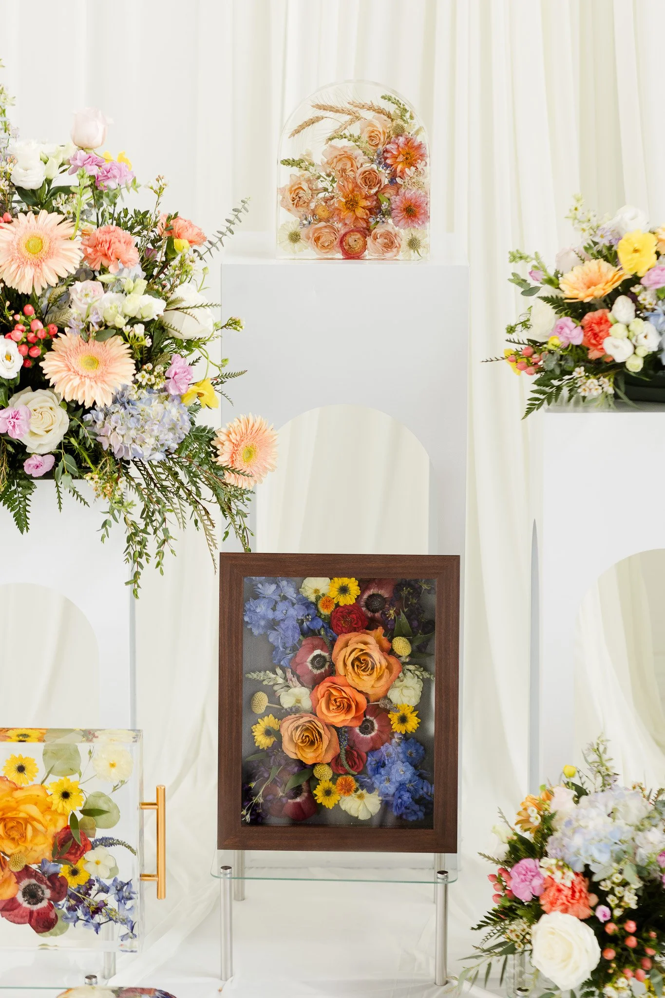 Display of various framed and glass-encased floral arrangements with pink, orange, purple, and white flowers on white backgrounds.
