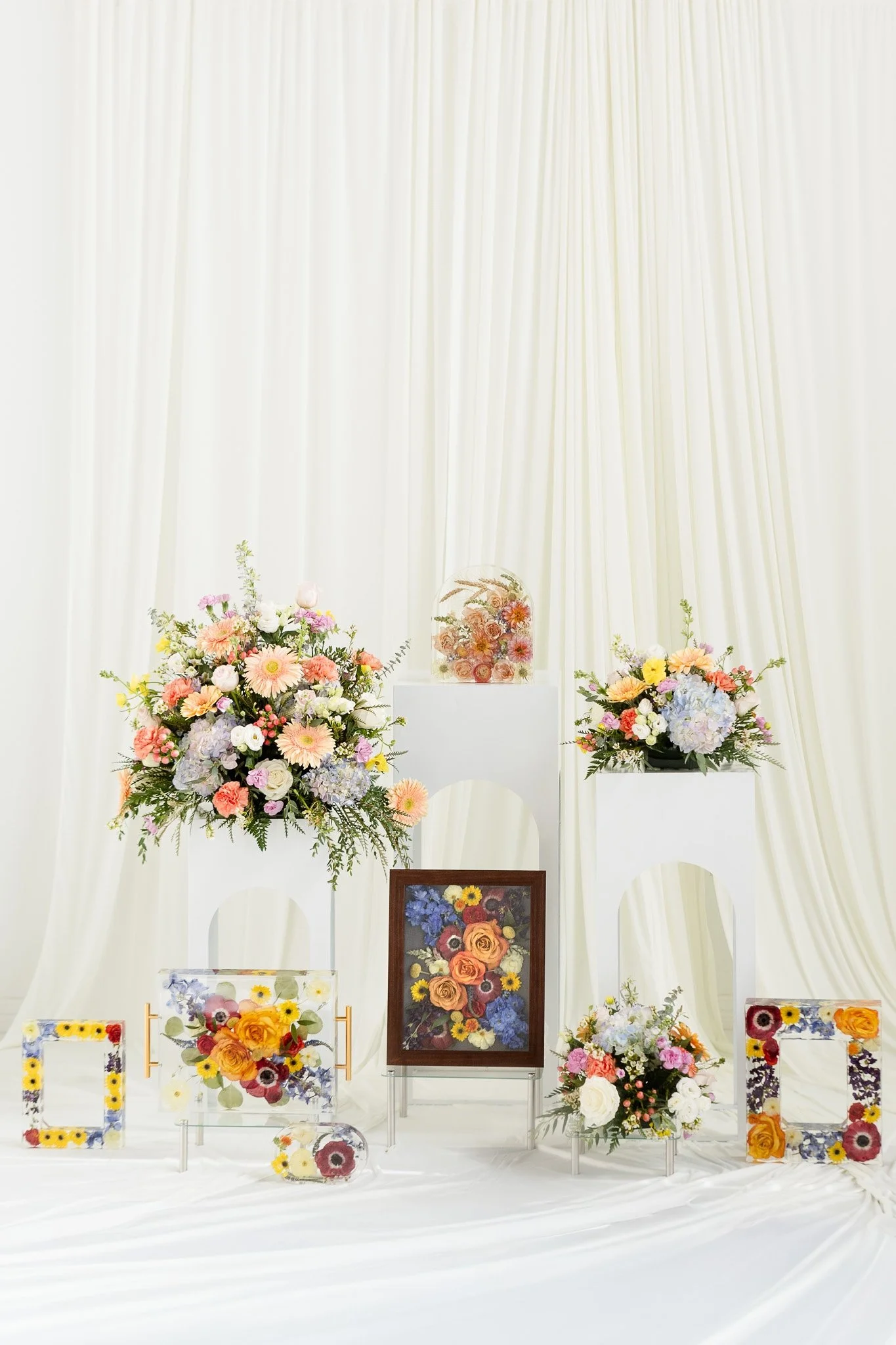Various floral arrangements and framed floral artwork displayed on white pedestals and stands, set against a backdrop of white curtains.