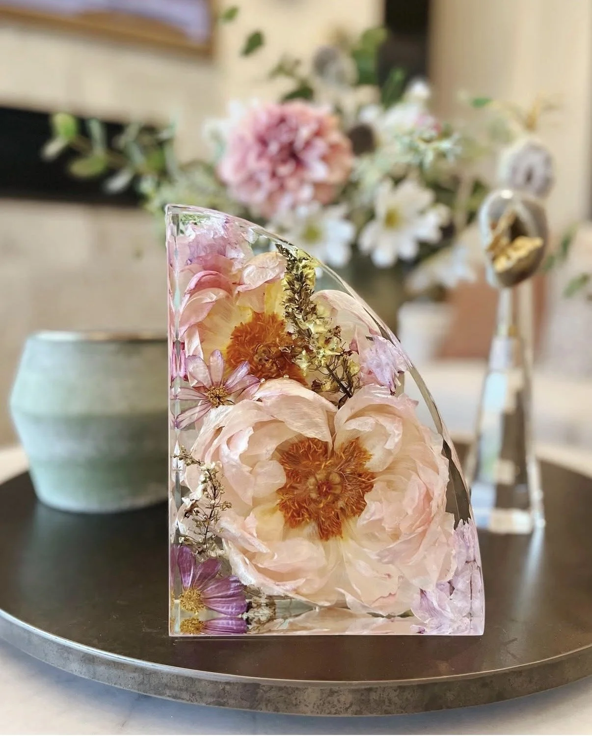 A glass art piece with embedded pink and orange flowers placed on a black tray in front of a blurred background of a flower arrangement and a ceramic pot.