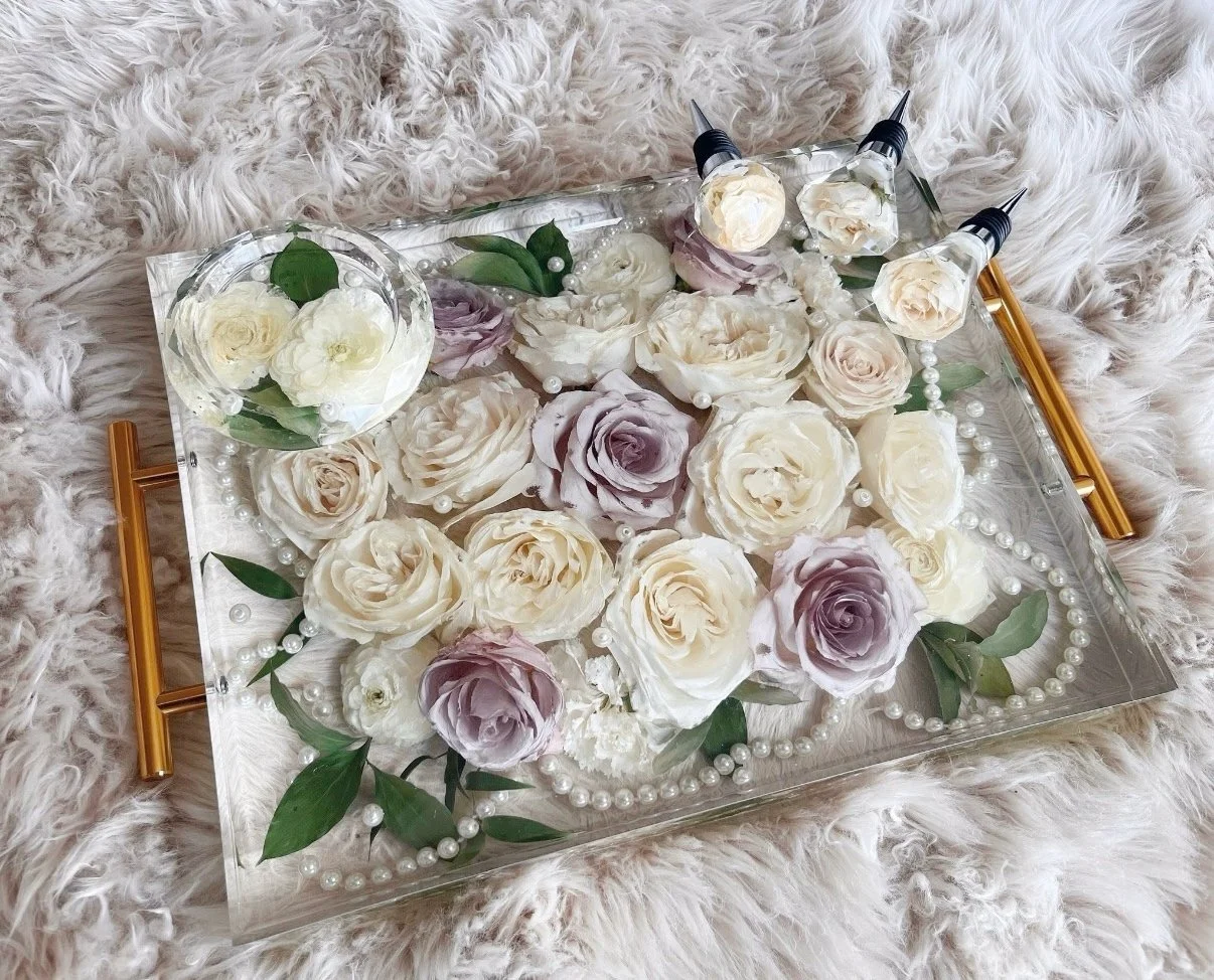 A rectangular transparent tray with gold handles, filled with white and lavender roses, pearl necklaces, and green leaves, resting on a fluffy cream-colored surface.