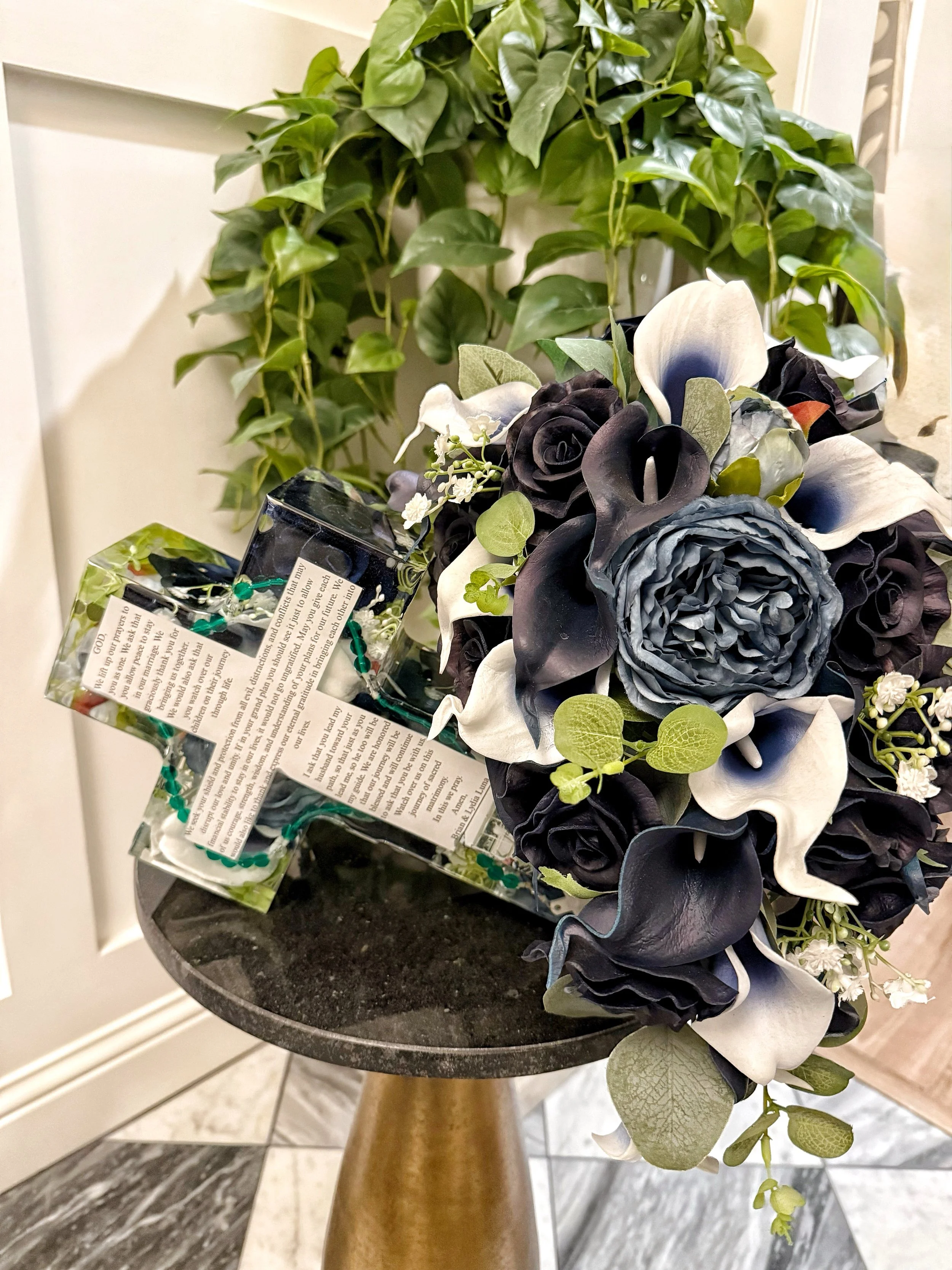 A funeral arrangement with black and white flowers, a small cross, and prayer cards, placed on a black round table with a gold base, in front of a leafy green plant.