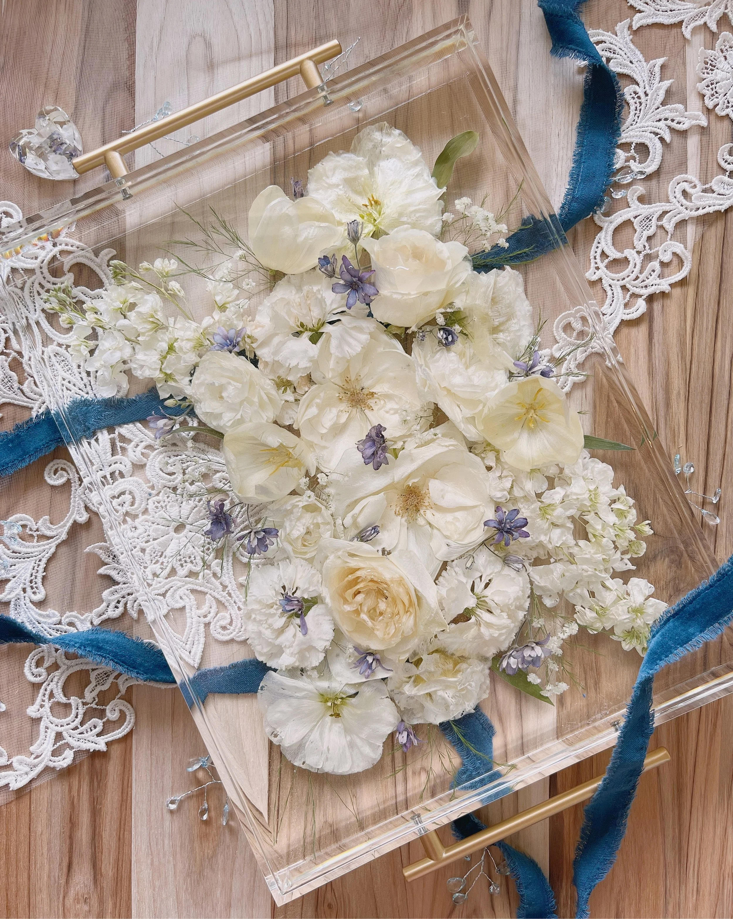 A floral arrangement inside a transparent box with white and pale yellow flowers, small purple blooms, and greenery, decorated with blue ribbons and lace on a wooden surface.