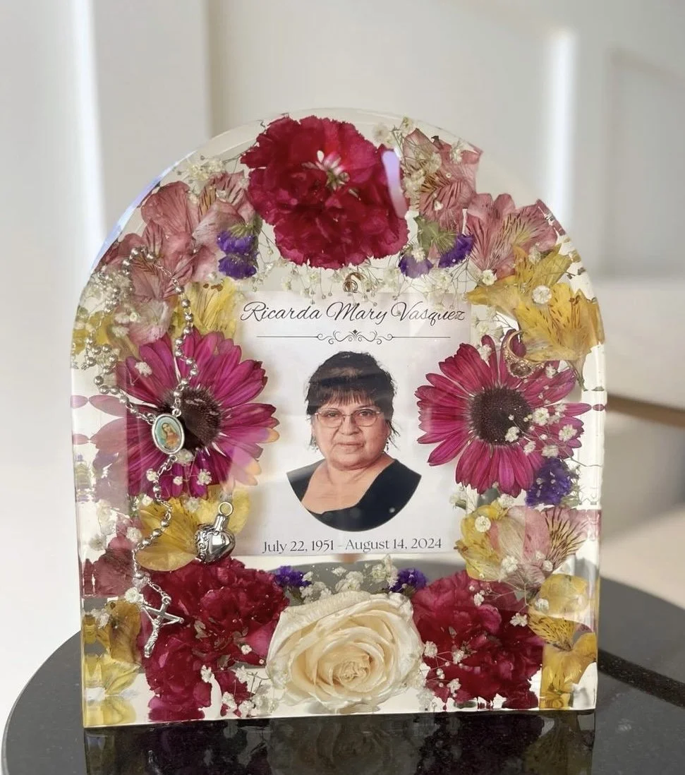 A memorial glass plaque with a portrait of a woman, surrounded by pressed flowers in pink, purple, yellow, and white, and some jewelry. The plaque bears the name Ricarda Mary Vasquez and her birth and death dates, July 22, 1951, and August 14, 2024.