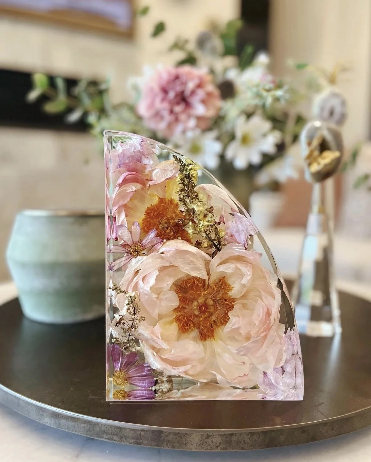 A half bookend resin block with embedded pink and orange flowers, placed on a round black tray in front of a blurred bouquet of flowers and a ceramic bowl.