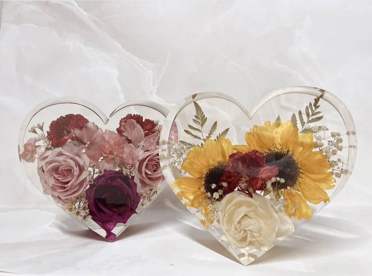 Two transparent heart-shaped glass photo frames filled with preserved flowers. The left frame contains pink roses and red carnations, while the right frame features yellow sunflowers, white roses, and small white filler flowers. Both frames are decor