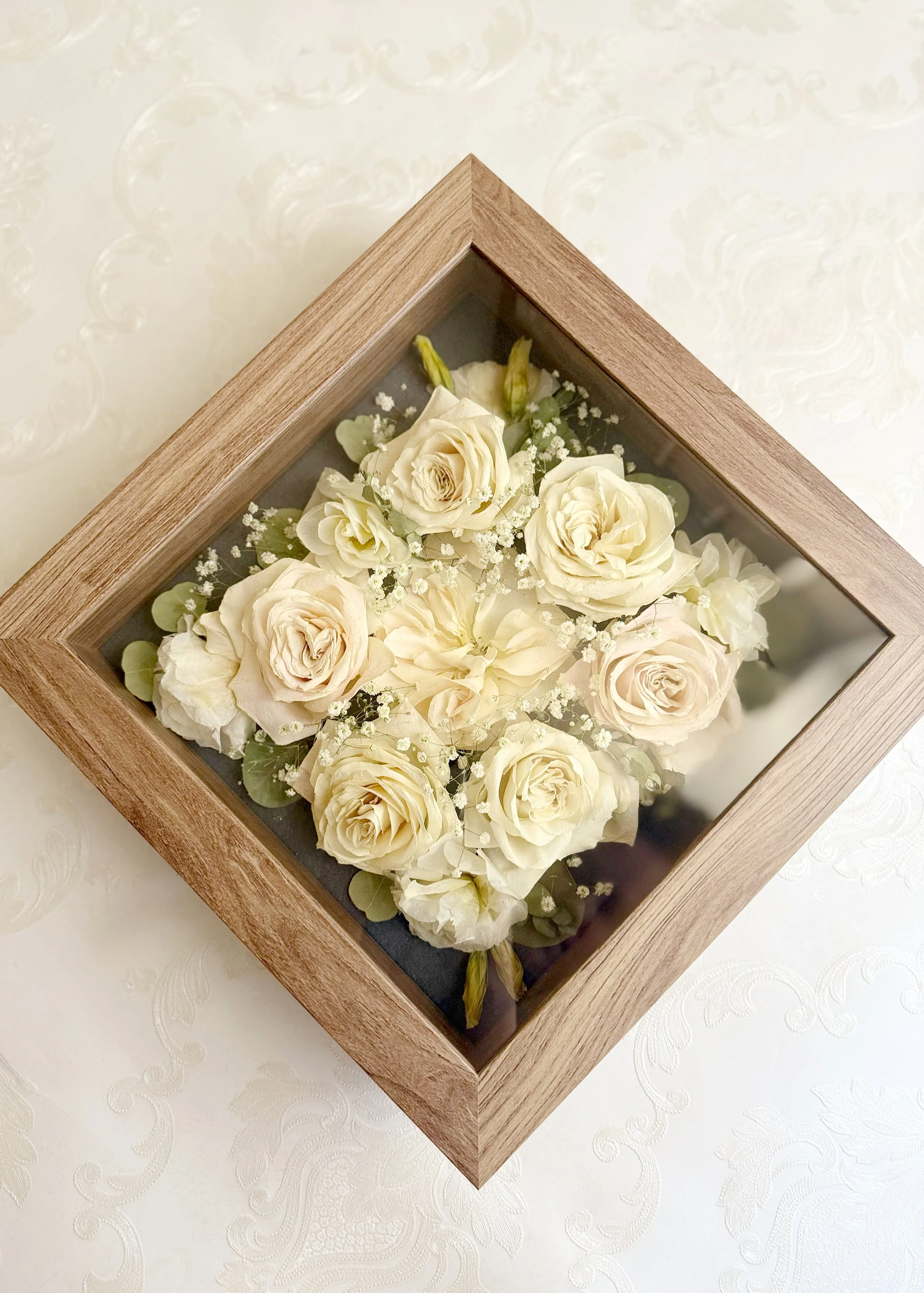 A display of white roses and small white filler flowers in a wooden frame with glass cover.