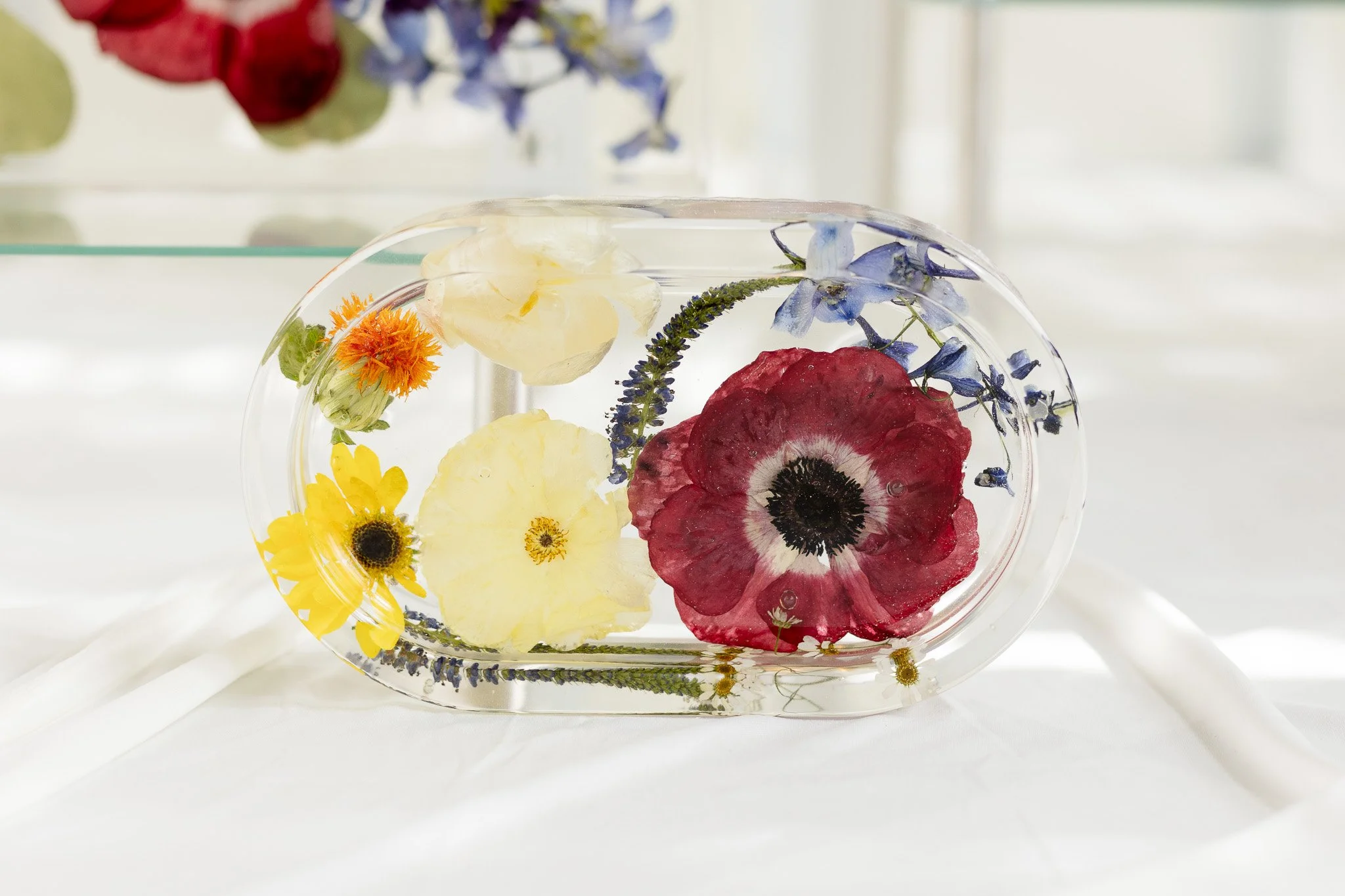 A resin oval-shaped mold containing floating colorful flowers including red, yellow, purple, and orange blossoms with green stems, set against a blurred background with natural light.