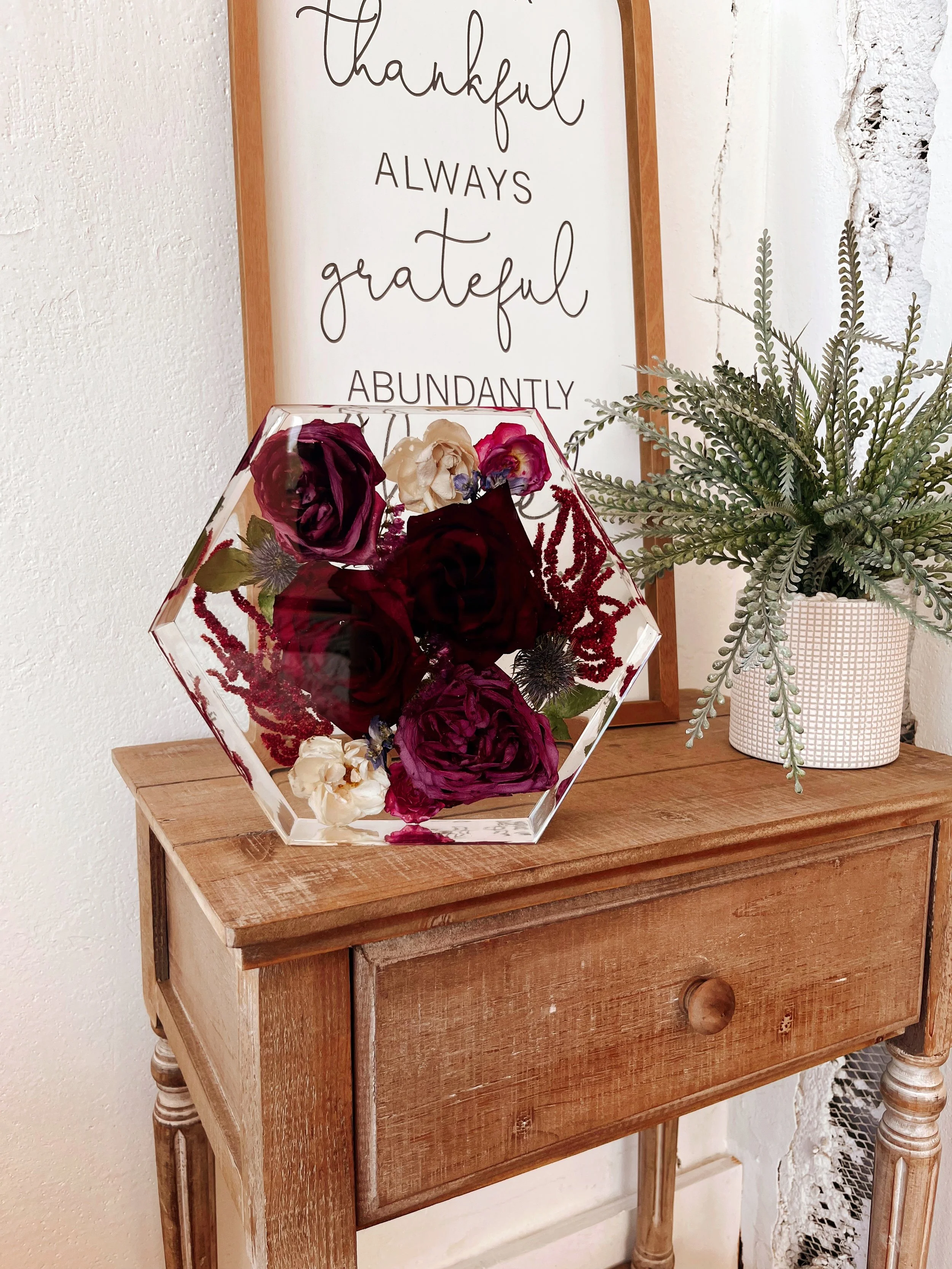 Decorative floral arrangement in a geometric glass vase with red and white flowers on a wooden table, next to a potted plant with green foliage, and a framed quote on the wall that reads "thankful, always grateful, abundantly".