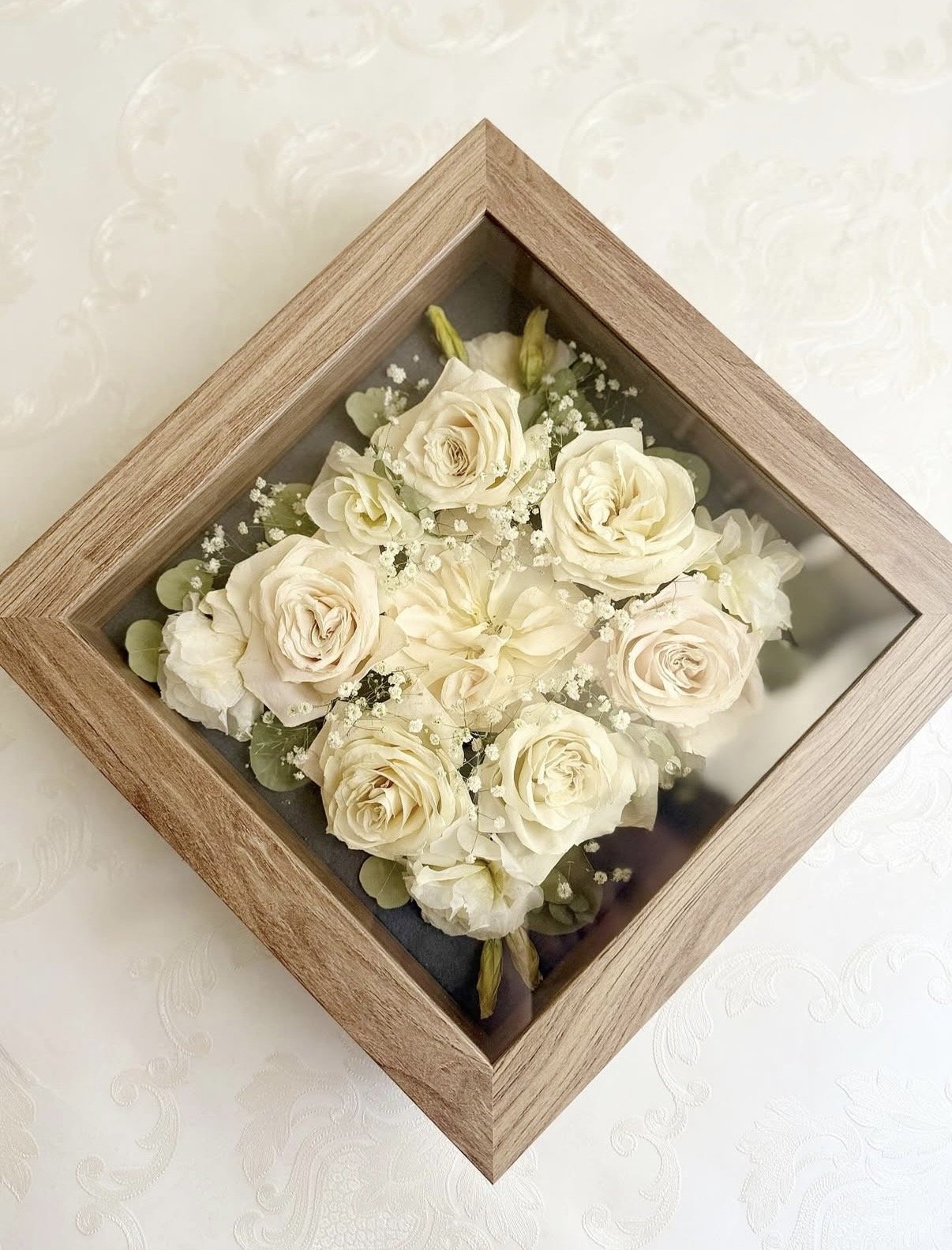 A wooden shadow box frame with glass top containing a bouquet of white roses and small white filler flowers arranged inside.