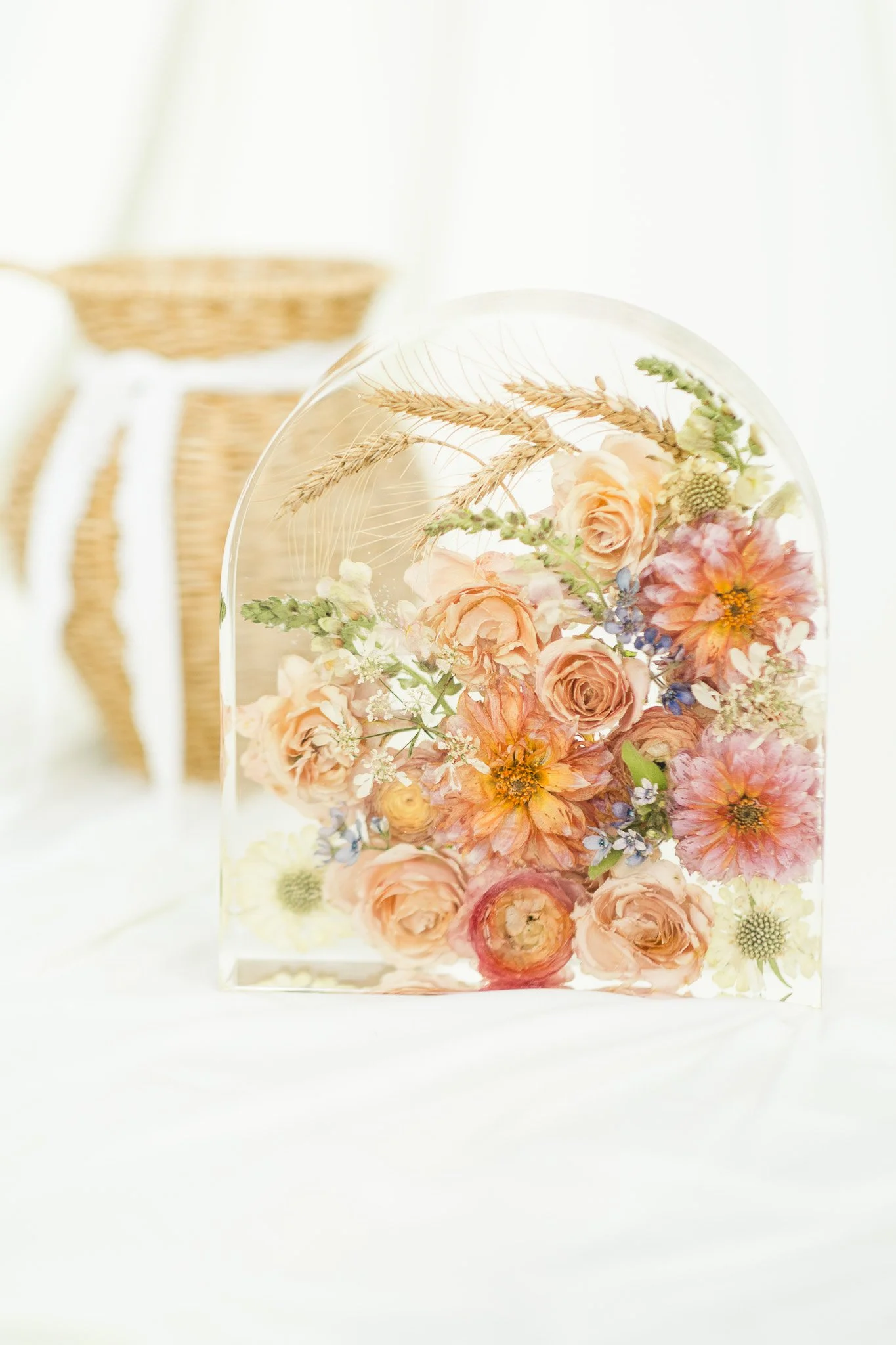 A glass dome filled with floating pastel-colored flowers and wheat stalks, with a blurred wicker basket in the background.