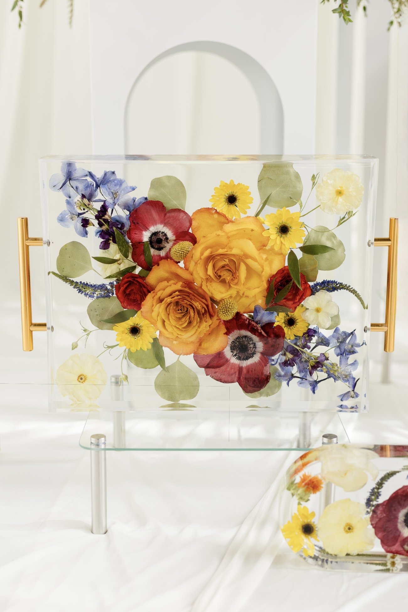 A resin tray display case containing a colorful arrangement of flowers, including yellow roses, red poppies, purple delphiniums, yellow daisies, and white flowers, with green leaves, set on a clear glass shelf.