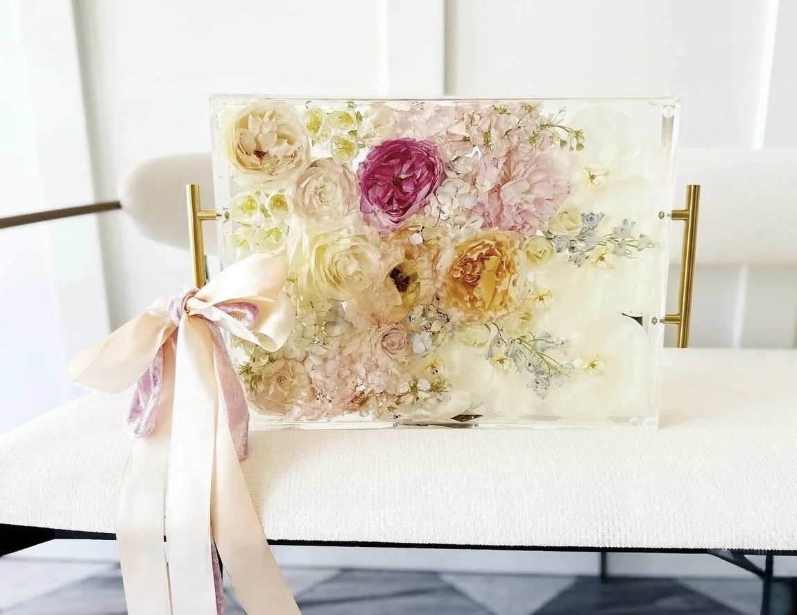 A rectangular clear acrylic resin tray filled with various pink, white, cream, and peach flowers, decorated with a large satin bow in shades of pink and ivory, placed on a white tablecloth.
