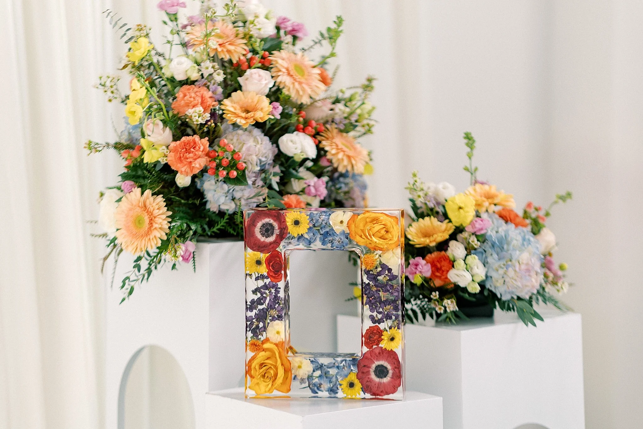 Floral arrangement of mixed flowers including pink, yellow, orange, and purple blooms on white pedestals, with a rectangular glass frame decorated with similar flowers in front of the arrangements.