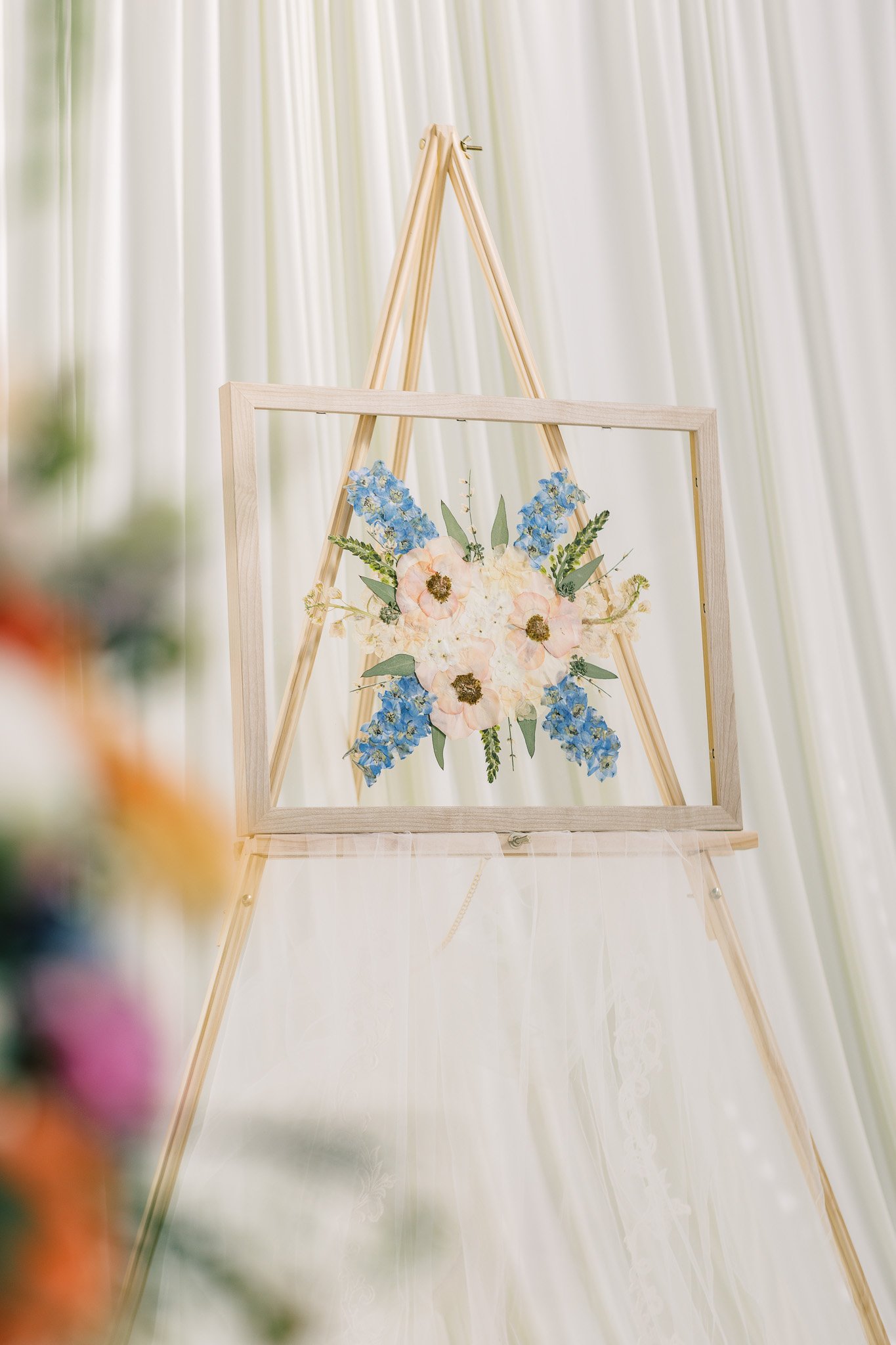 Floral artwork of pink, white, and blue flowers inside a wooden frame on a gold easel, with white curtains in the background.